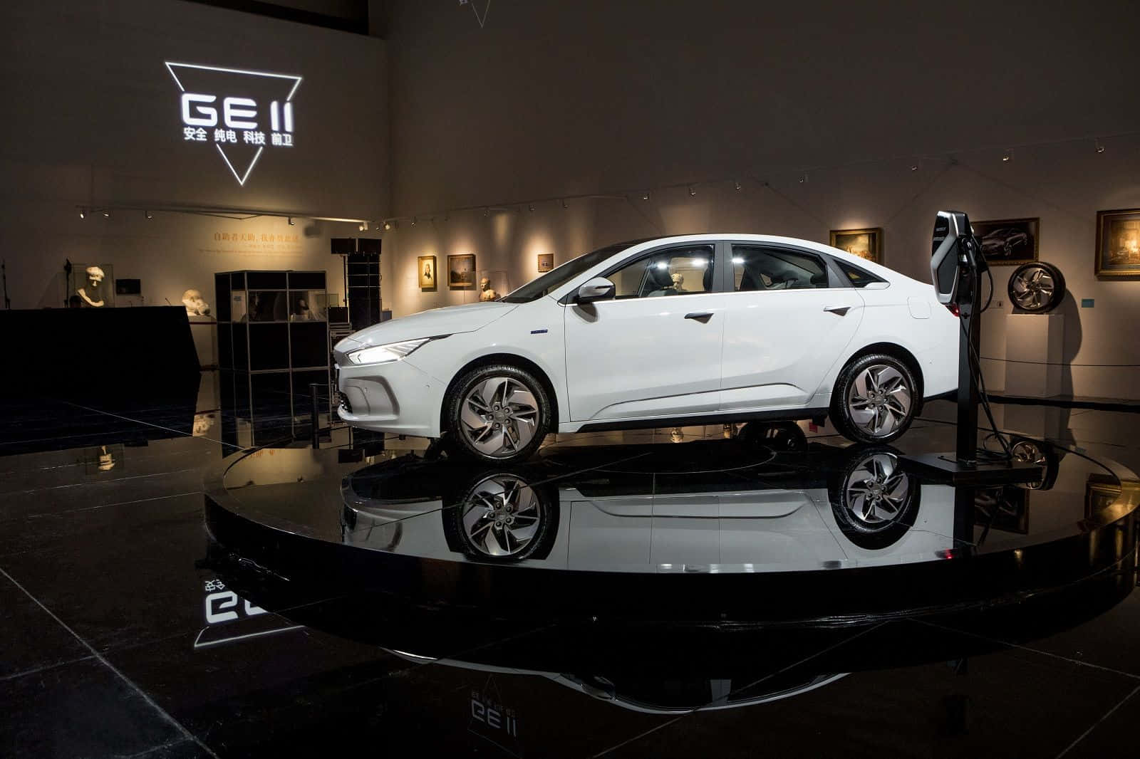 Geely 桌布 壁紙 — 一架時尚的白色電動轎車陳列在光亮的黑色平台上，位於燈光昏暗的現代展廳內，配有反光表面和品牌燈光。