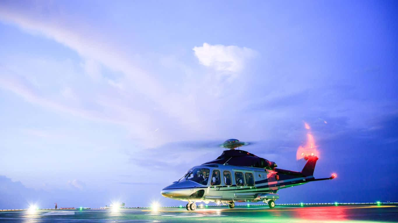 1366x768 Helicopters 桌布 壁紙 — 一架時尚的直升機在暮色天空和柔軟雲朵下，降落在燈光明亮的直升機停機坪上。