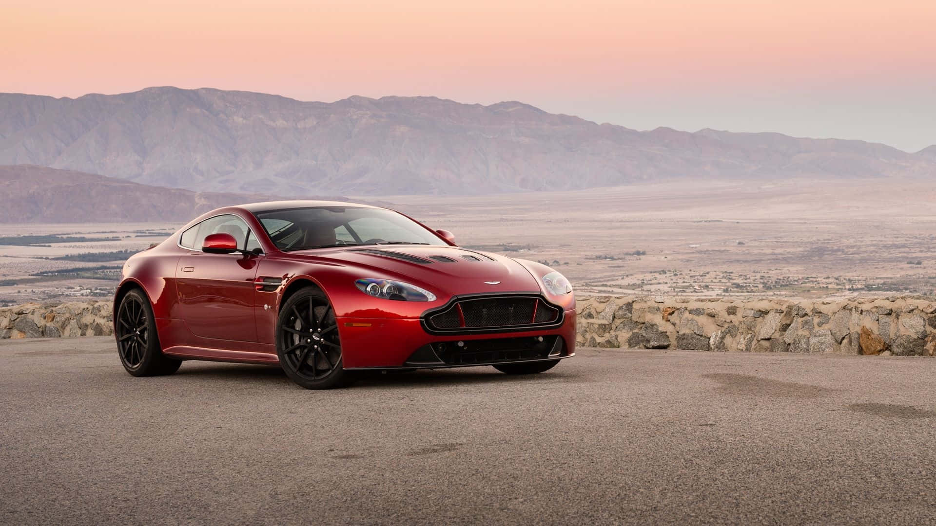 Aston Martin V12 Vantage 桌布 壁紙 — 一架時尚的紅色Aston Martin跑車停放在鋪砌的觀景點上，背景是山脈和粉紅色的日落。
