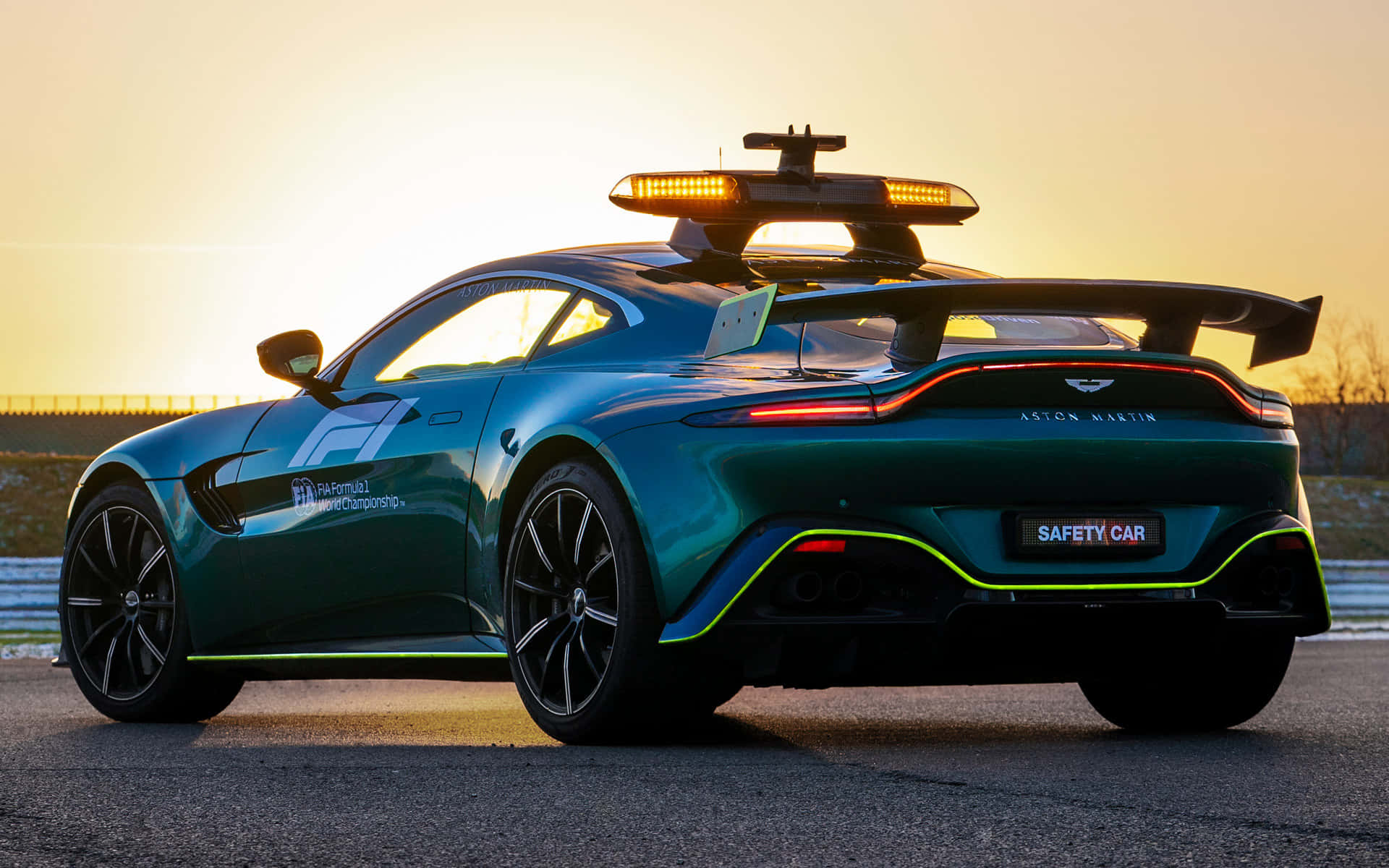 奧斯頓馬丁 桌布 壁紙 — 一架時尚的綠色 Aston Martin 安全車，閃爍著燈光，停泊在金色日落下的賽道上。
