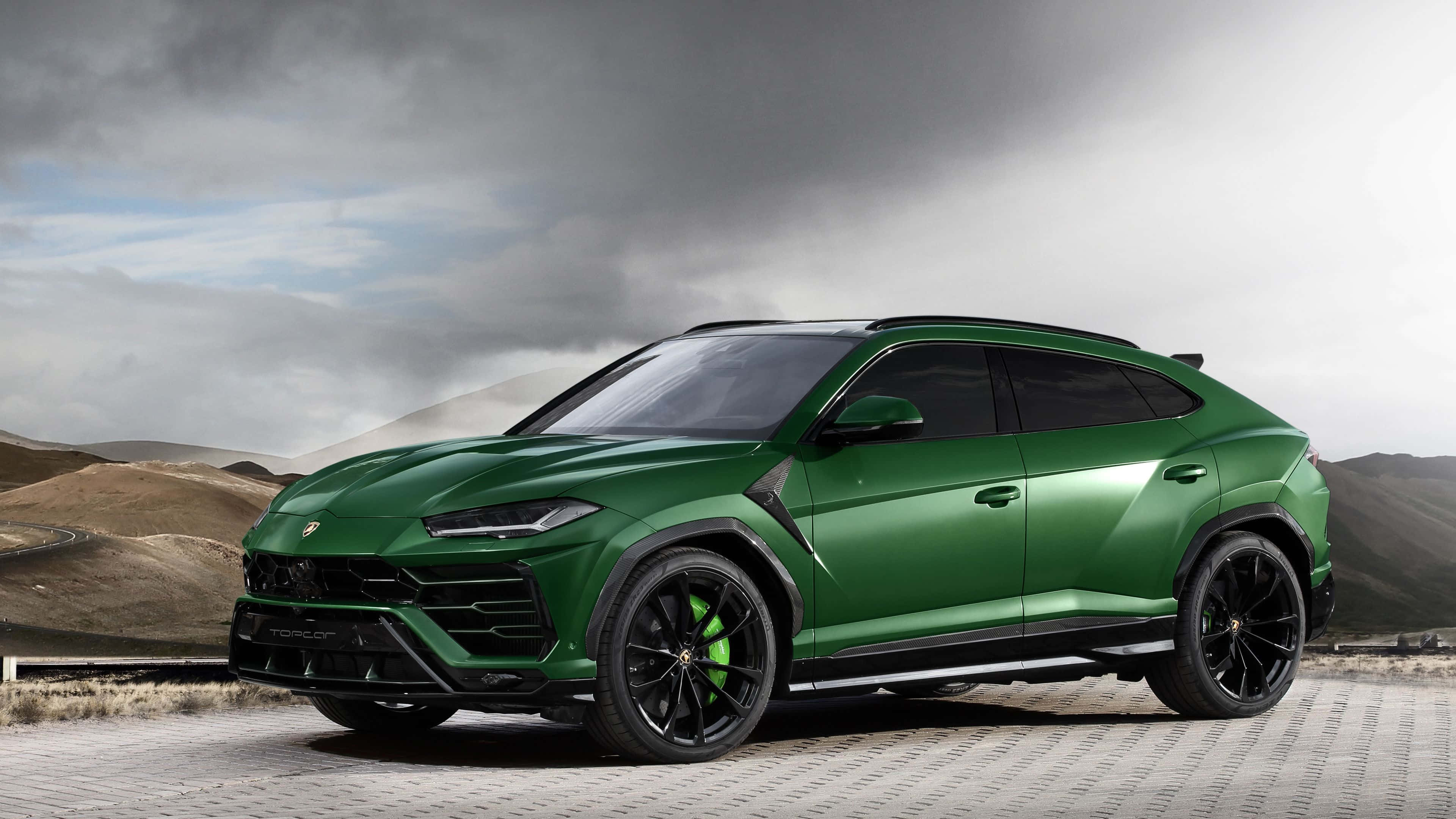 Lamborghini Urus 桌布 壁紙 — 一架時尚的綠色藍寶堅尼 Urus SUV 停放在鋪砌道路上，背景是戲劇性的多雲天空和連綿起伏的丘陵。