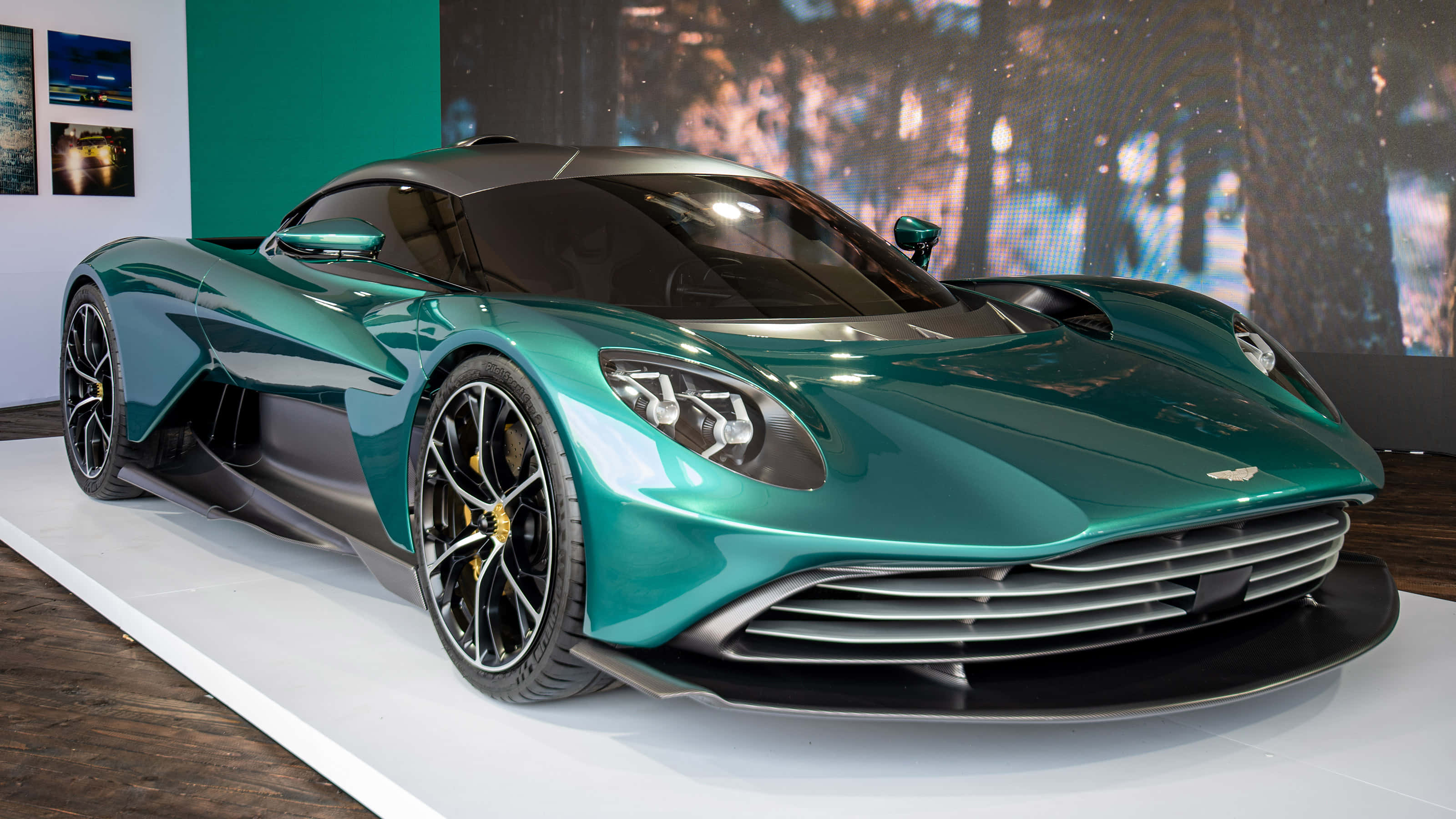 奧斯頓馬丁 桌布 壁紙 — 一架時尚的藍綠色 Aston Martin 超級跑車在現代展廳的白色展示台上，在明亮燈光下閃閃發光。
