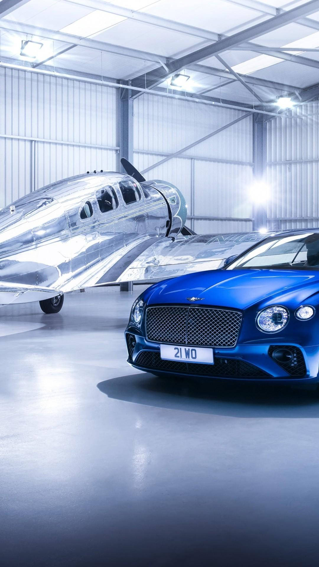 手機 桌布 壁紙 — 一架時尚的藍色 Bentley 跑車和一架閃亮的銀色復古飛機並排展示在明亮的工業機庫中。