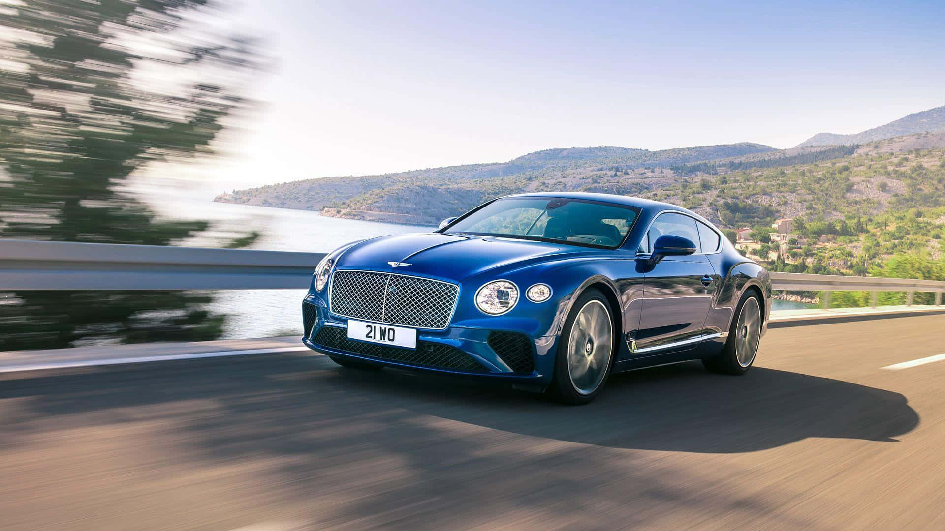 Convertible 桌布 壁紙 — 一架時尚的藍色 Bentley Continental GT 沿著海岸高速公路飛馳，背景是模糊的綠色山丘和水面。