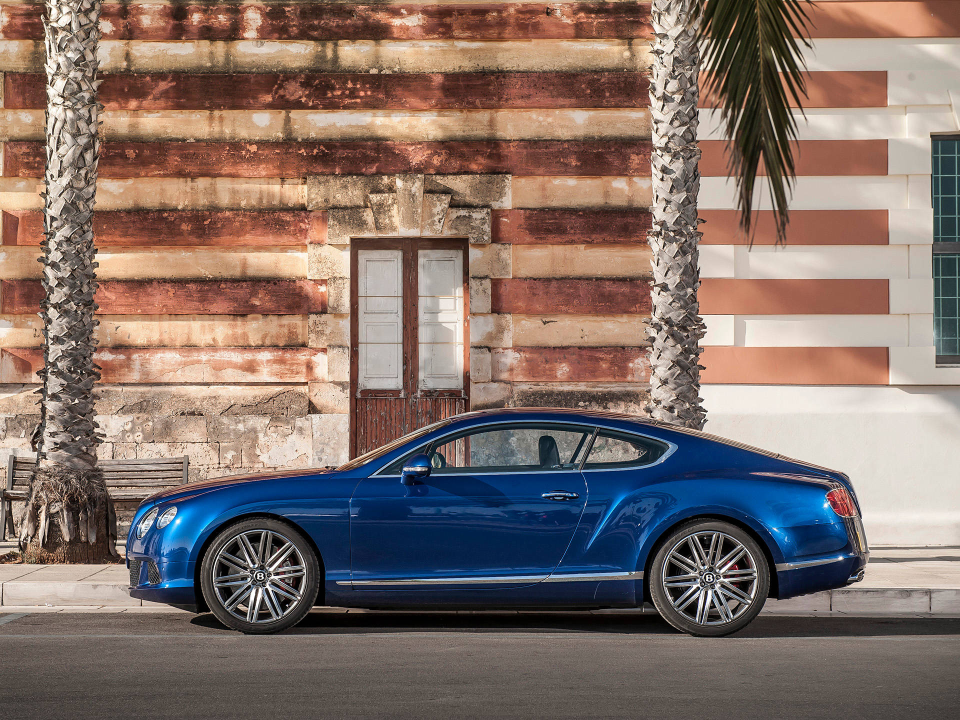 bentley HD 桌布 壁紙 — 一架時尚的藍色 Bentley Continental GT 停泊在棕櫚樹旁，背景是風化的條紋建築外牆。