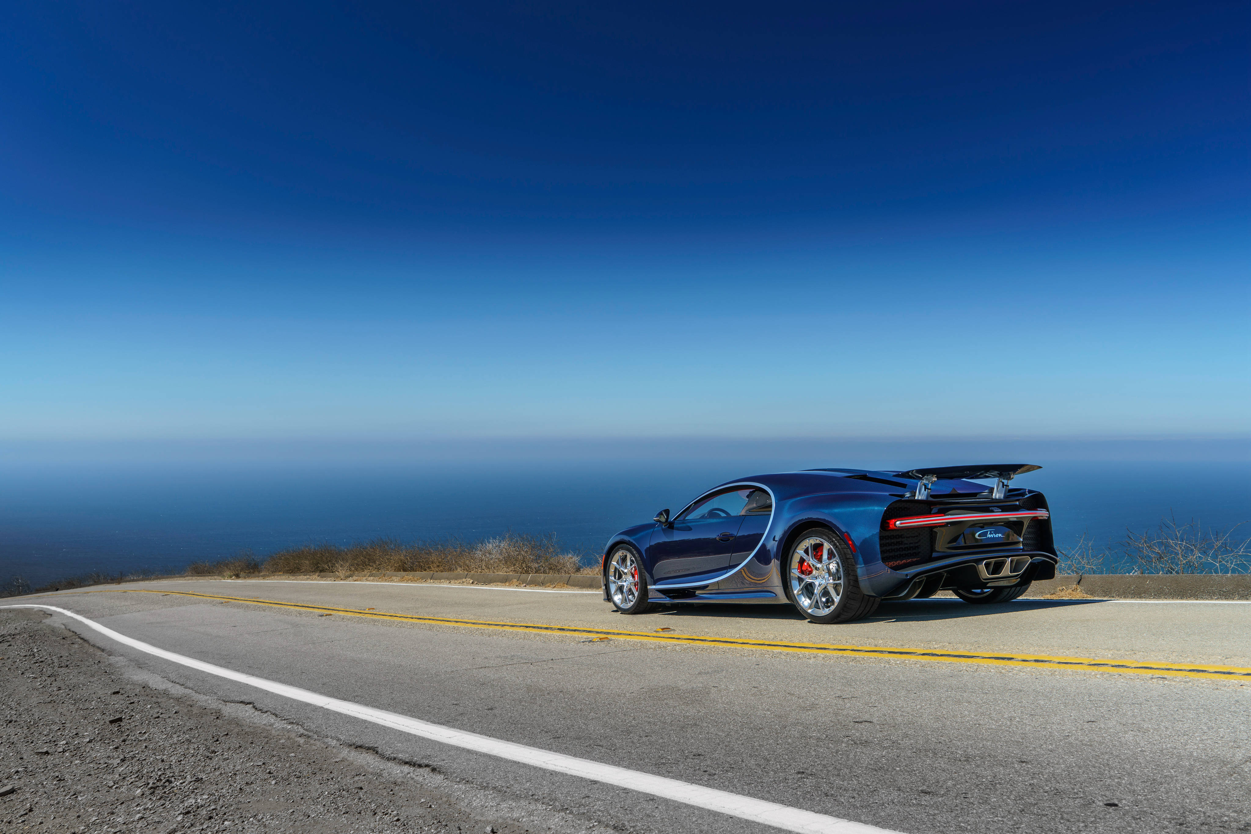 布加迪 桌布 壁紙 — 一架時尚的藍色 Bugatti Chiron 停泊在沿海懸崖公路上，俯瞰著清澈藍天下的海洋。