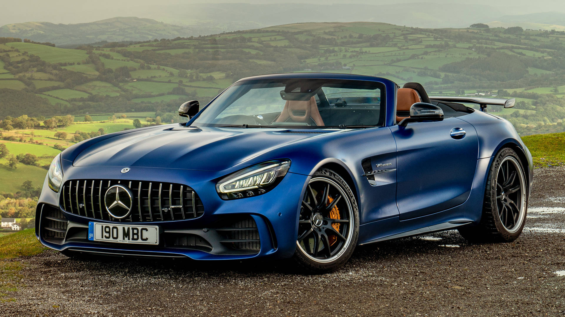 Gtr 桌布 壁紙 — 一架時尚的藍色 Mercedes-AMG GT Roadster 停在碎石路上，俯瞰著連綿起伏的綠色山丘，在柔和的光線下。