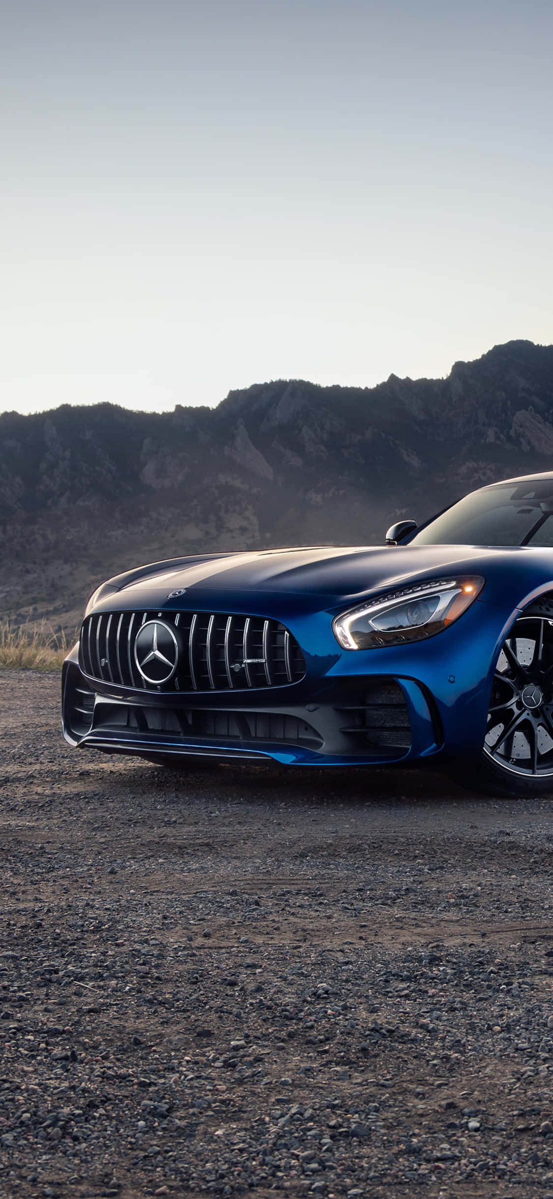 iPhone X Amg Gt-r 桌布 壁紙 — 一架時尚的藍色 Mercedes-AMG GT 停泊在碎石上，背景是崎嶇的山脈，天空柔和。