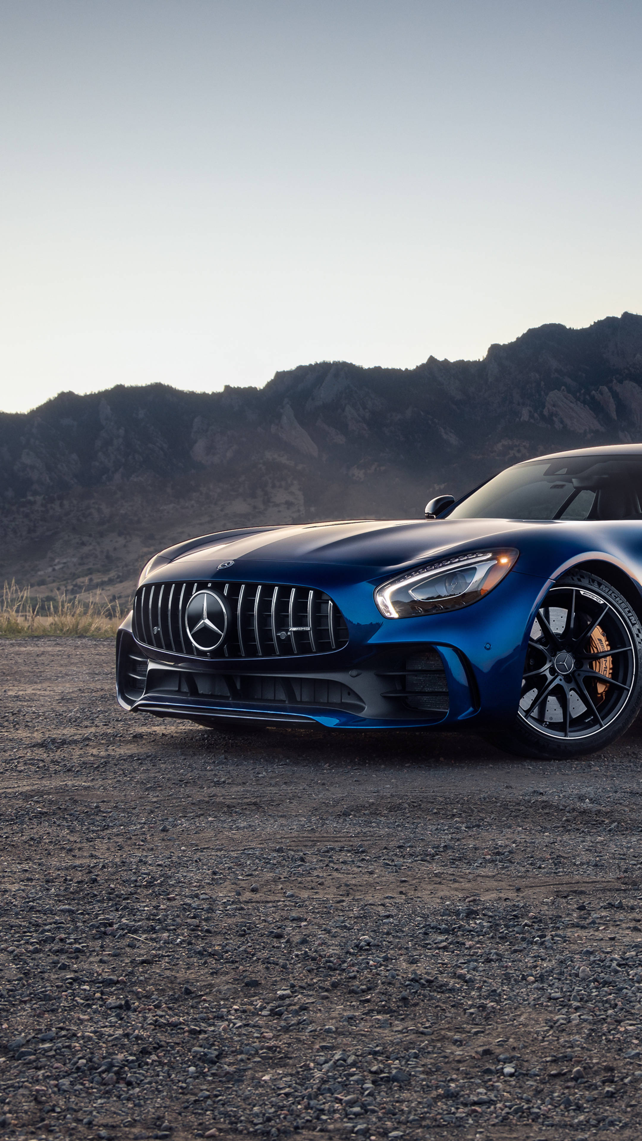 賓士 iPhone 桌布 壁紙 — 一架時尚的藍色 Mercedes-AMG GT 停泊在碎石上，背景是崎嶇的山脈，天空晴朗。