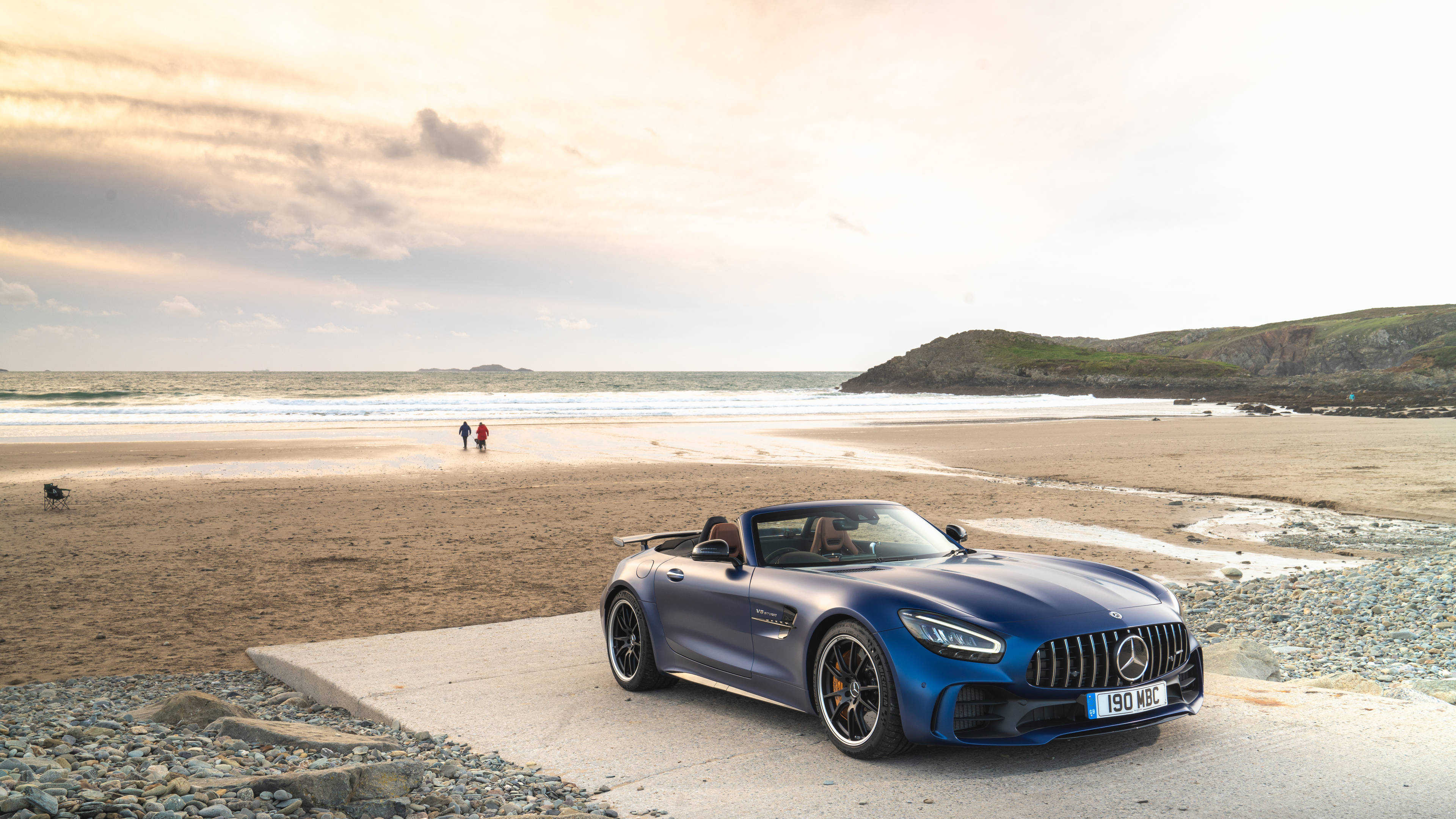 Gtr 桌布 壁紙 — 一架時尚的藍色 Mercedes-AMG GT Roadster 停泊在卵石海灘上，背景是海浪和遠處的懸崖，天空呈現柔和的金色。