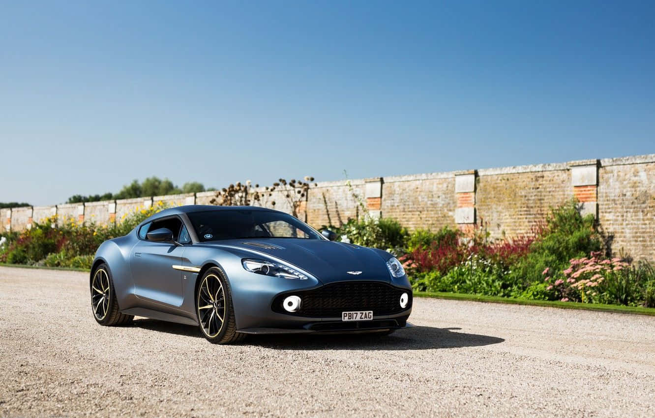 Aston Martin Vanquish 桌布 壁紙 — 一架時尚的藍色Aston Martin跑車停放在碎石車道上，旁邊是磚牆和花園，在晴朗的藍天下盛開著花朵。