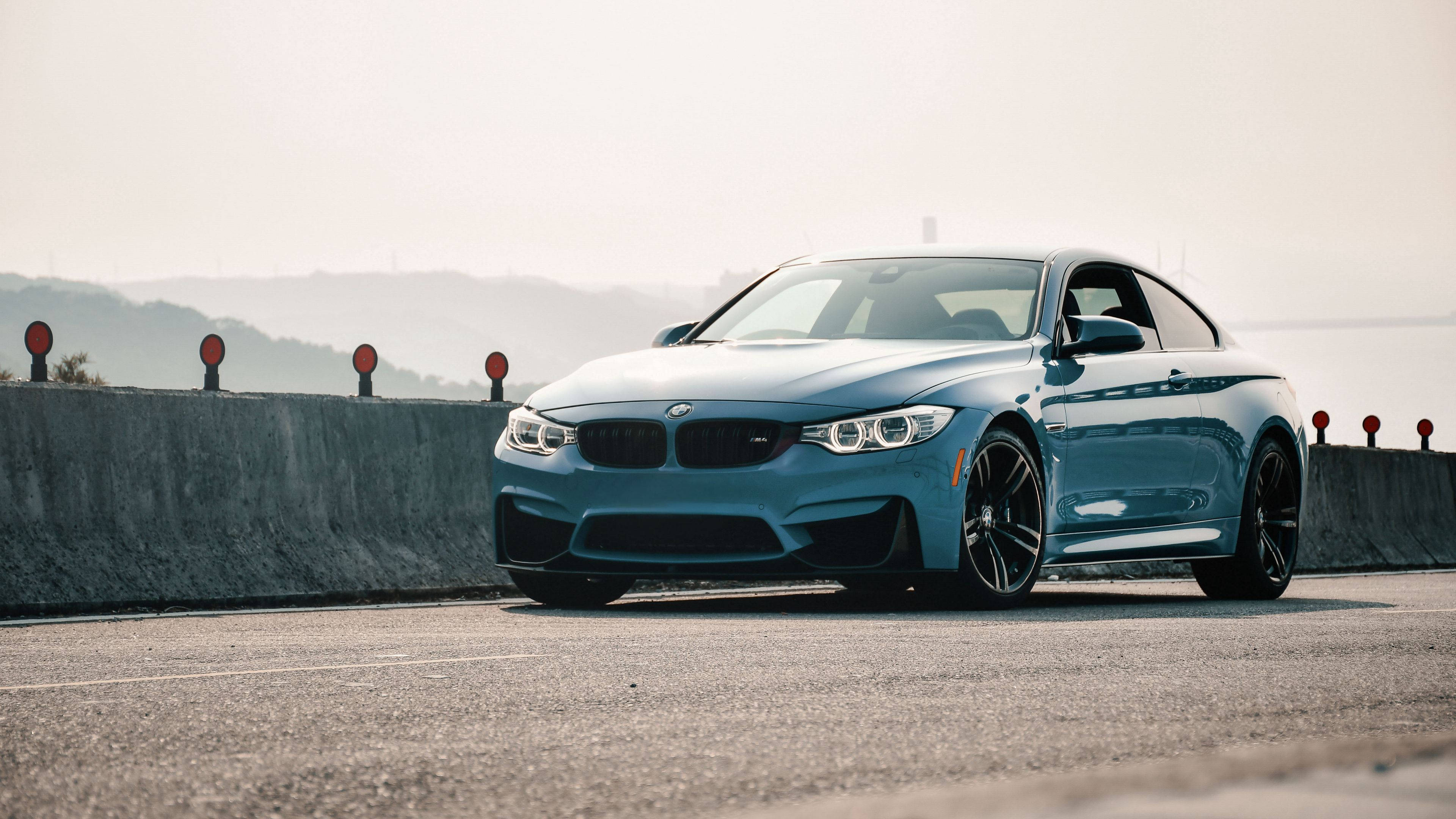4K BMW 壁紙 桌布 壁紙 — 一架時尚的藍色BMW M4轎跑車停泊在沿海公路上，背景是朦朧的山丘。