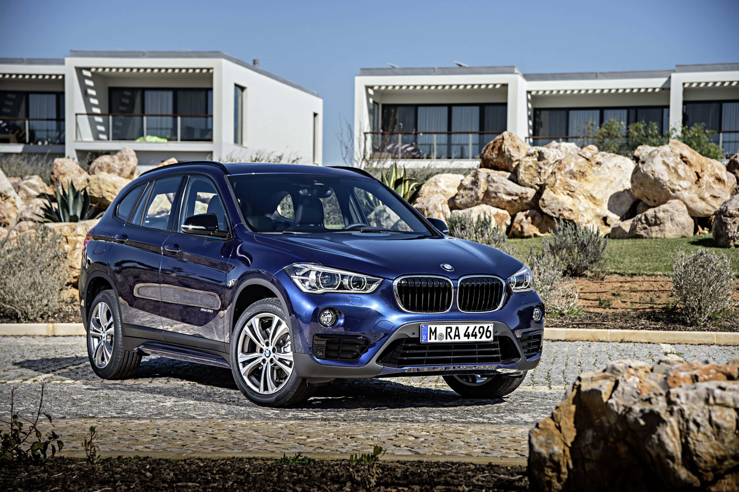 Bmw X1 桌布 壁紙 — 一架時尚的藍色BMW X1 SUV停泊在鵝卵石車道上，前方是現代白色別墅，周圍有大石頭和乾燥灌木，天空清澈湛藍。