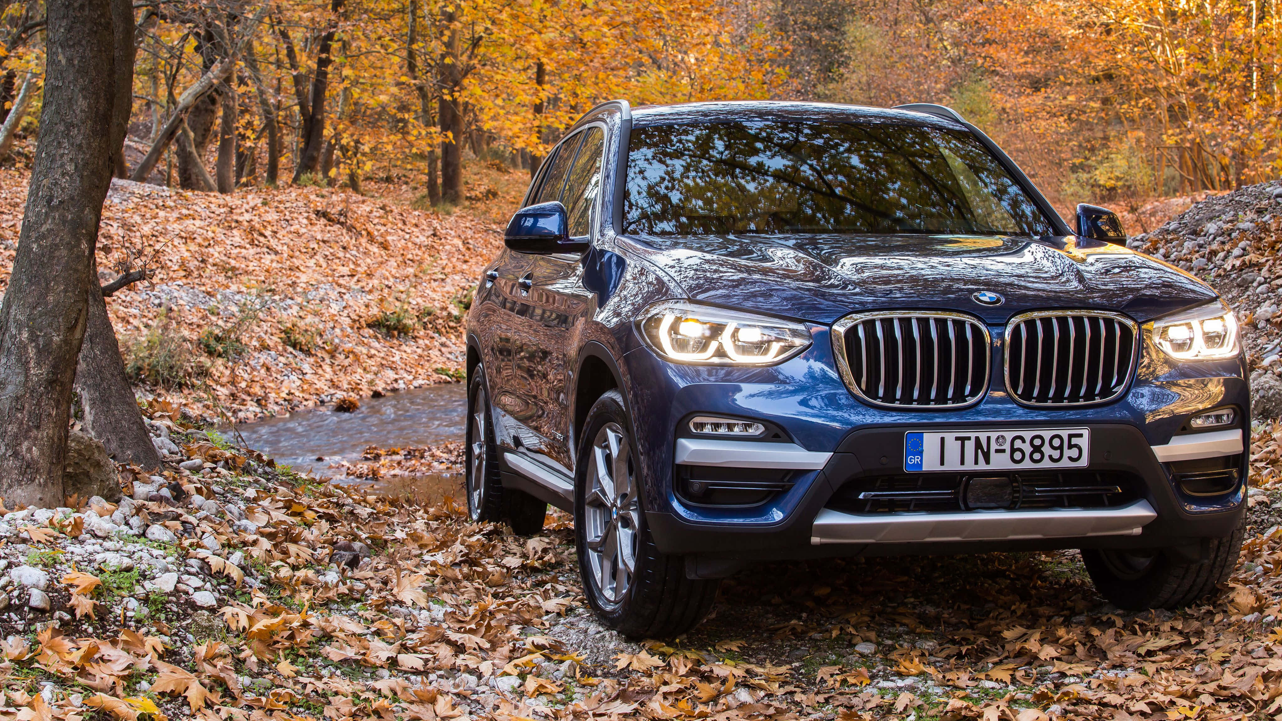 Bmw X3 桌布 壁紙 — 一架時尚的藍色BMW X3 SUV在柔和日光下行駛在鋪滿金色秋葉的森林中。
