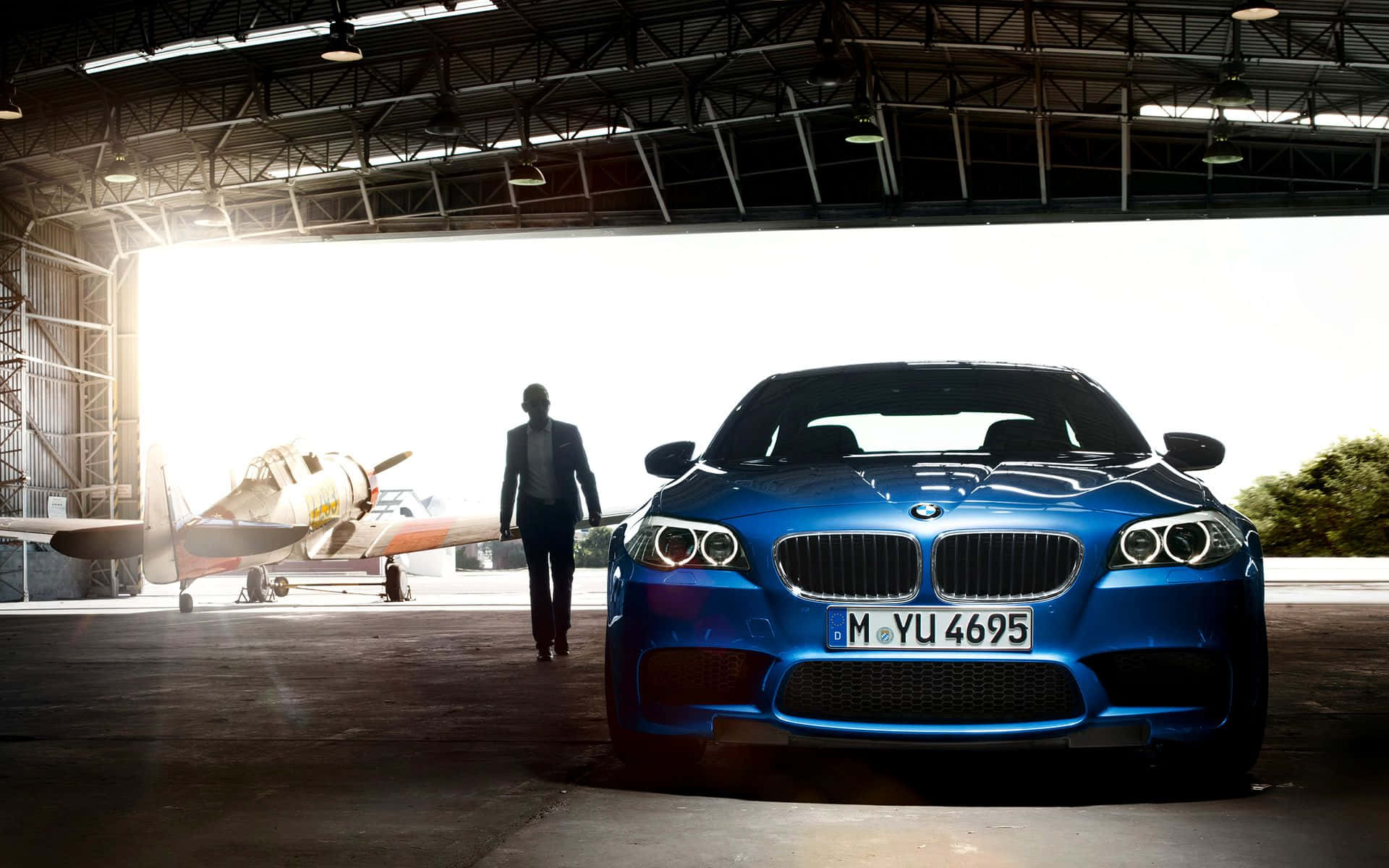 BMW 動圖 桌布 壁紙 — 一架時尚的藍色BMW跑車在機庫中閃閃發光，一名男子正穿過沐浴在明亮陽光下的復古飛機旁邊走向它。
