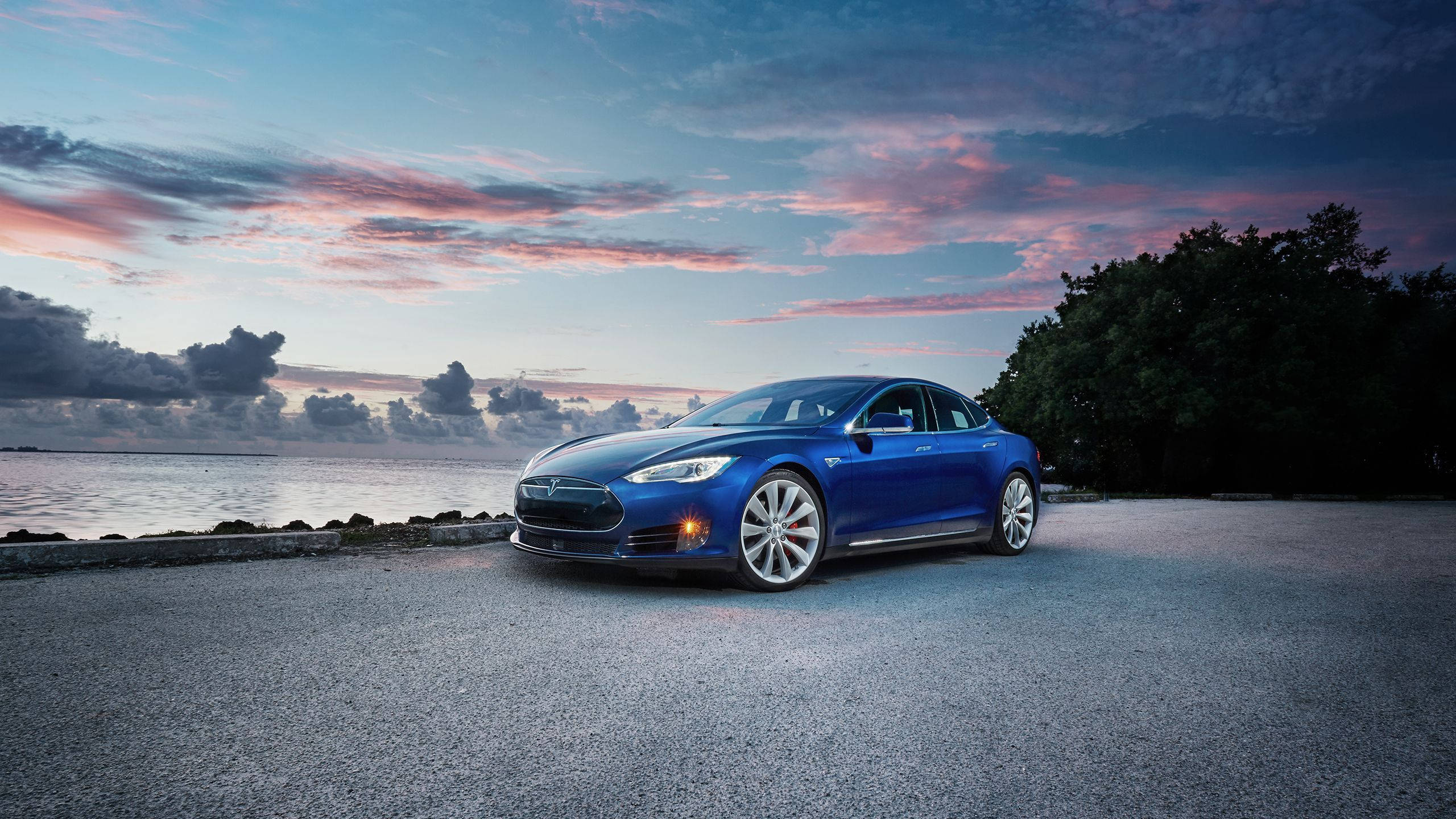 特斯拉 桌布 壁紙 — 一架時尚的藍色Tesla Model S停泊在黃昏的礫石岸邊，水面上方是充滿活力的粉紅色和紫色天空。