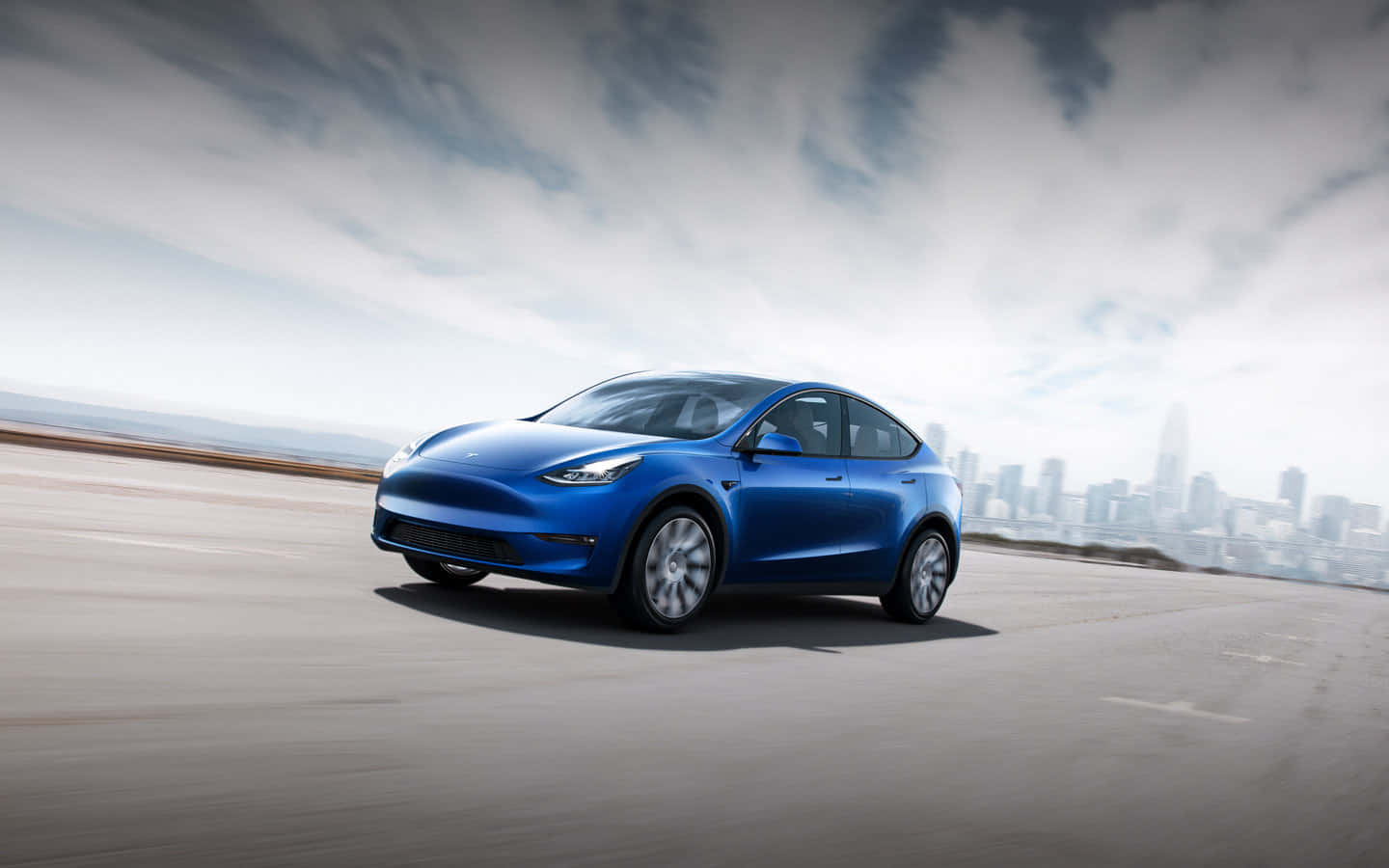 特斯拉 桌布 壁紙 — 一架時尚的藍色Tesla Model Y在沿海道路上行駛，遠處是朦朧的城市天際線，天空多雲。