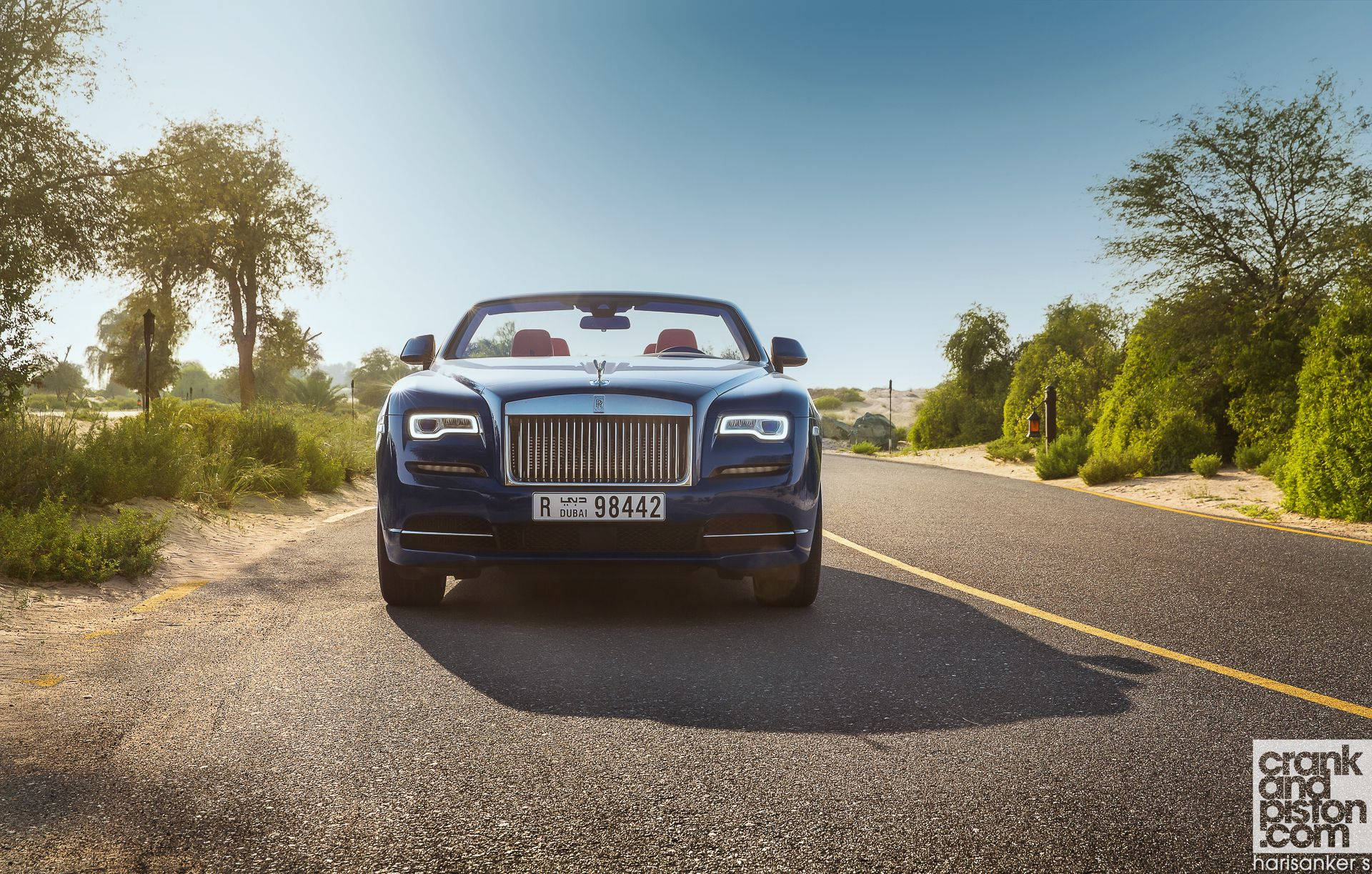Royce 桌布 壁紙 — 一架時尚的藍色勞斯萊斯敞篷車在陽光明媚的沙漠公路上向前行駛，兩旁是綠色灌木和樹木，天空湛藍清澈。