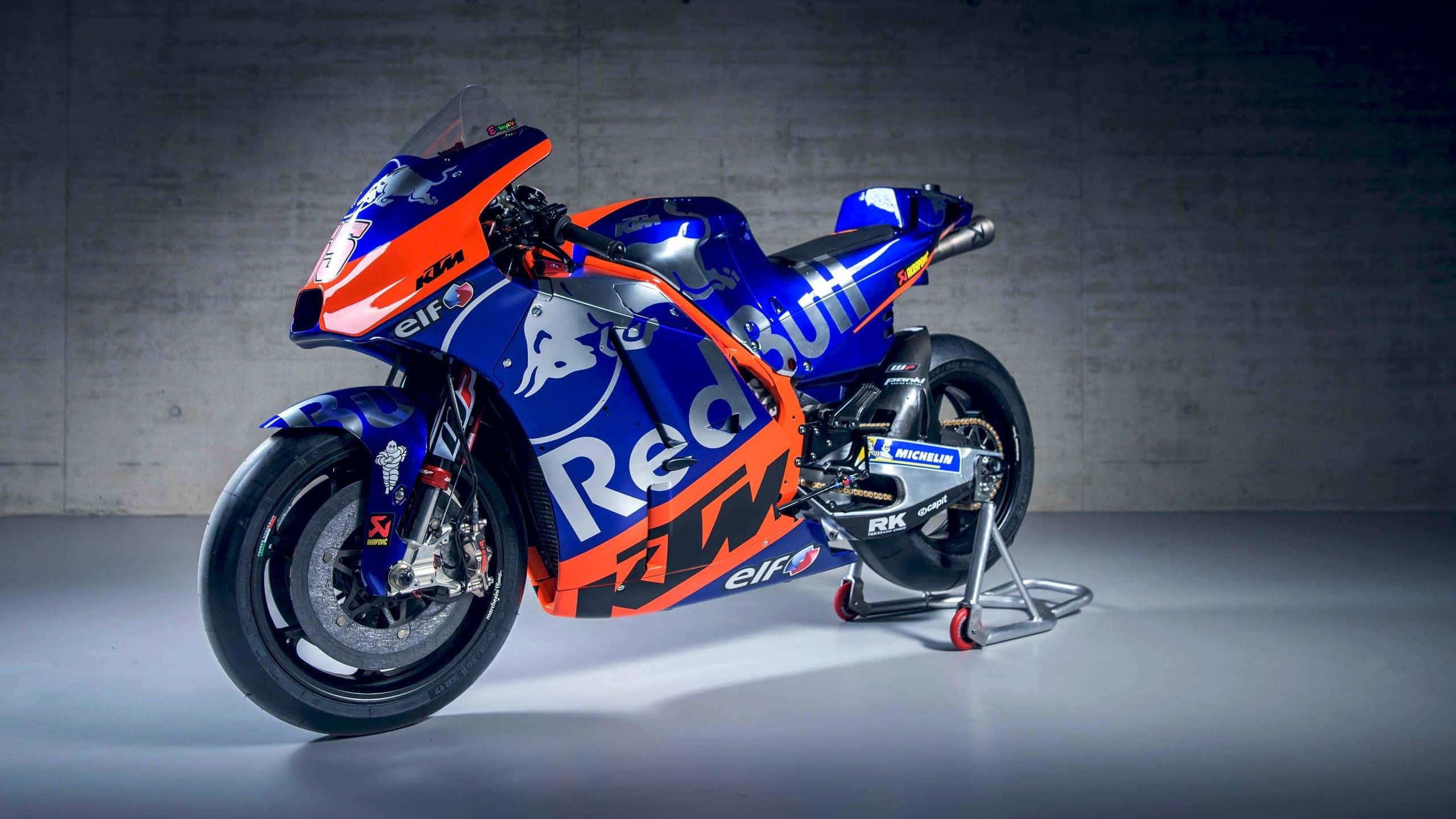 Ktm Bike 桌布 壁紙 — 一架時尚的藍色和橙色KTM Red Bull賽車摩托車，在簡約混凝土背景下立於支架上。