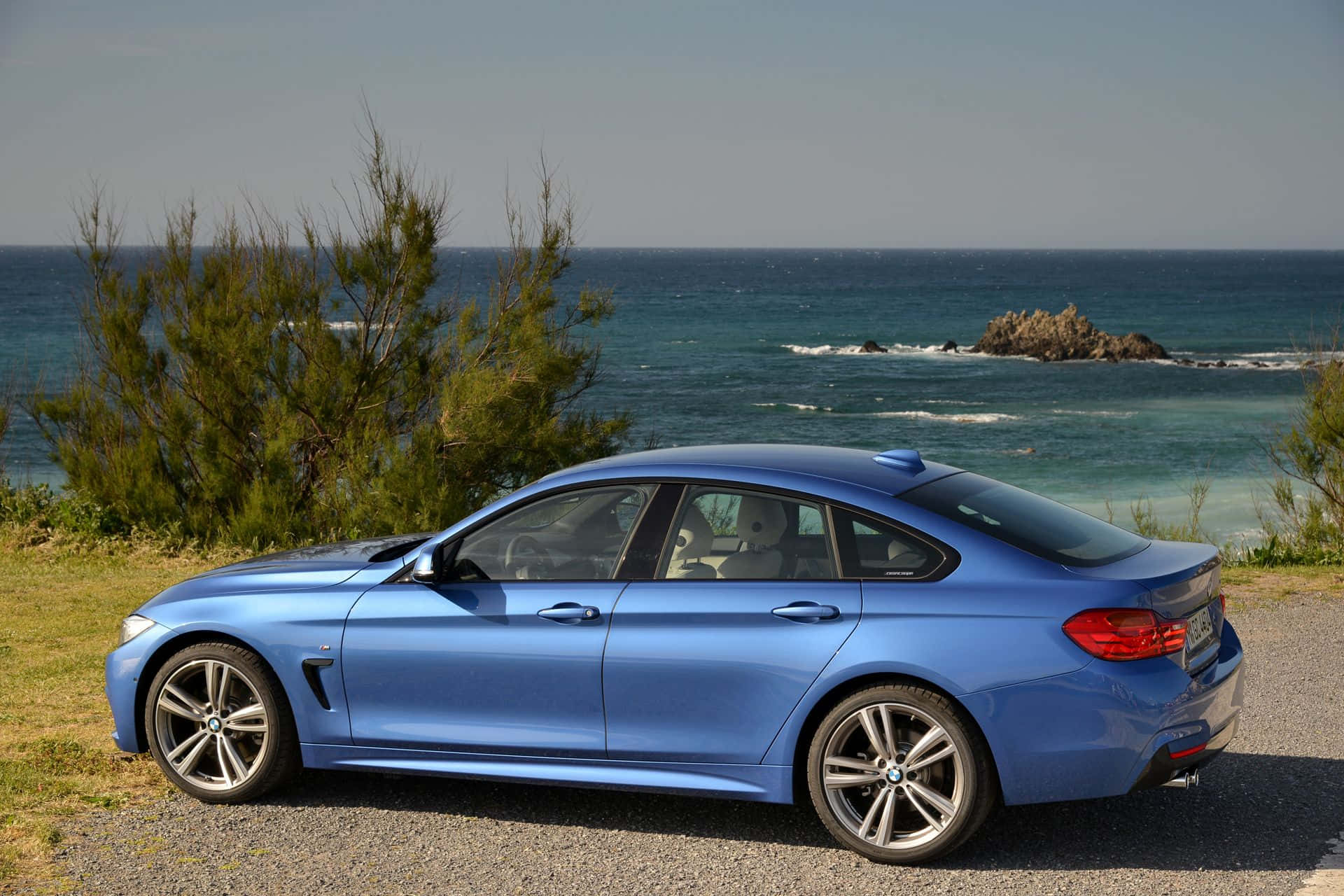 Bmw 4 Series 桌布 壁紙 — 一架時尚的藍色寶馬轎跑車停泊在海邊的碎石路上，背景有綠色灌木和碧綠的海水。