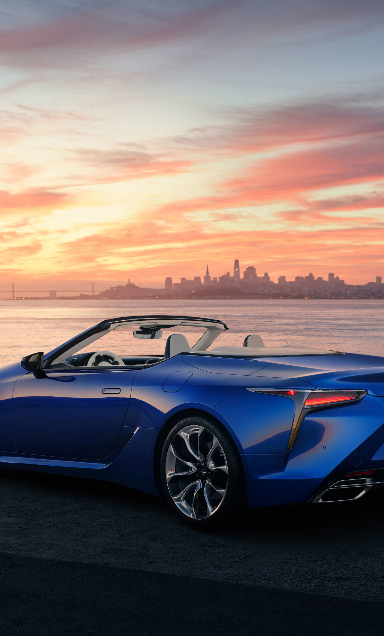 lexus iPhone 桌布 壁紙 — 一架時尚的藍色敞篷跑車停在水邊，夕陽西下，遠處是城市天際線。