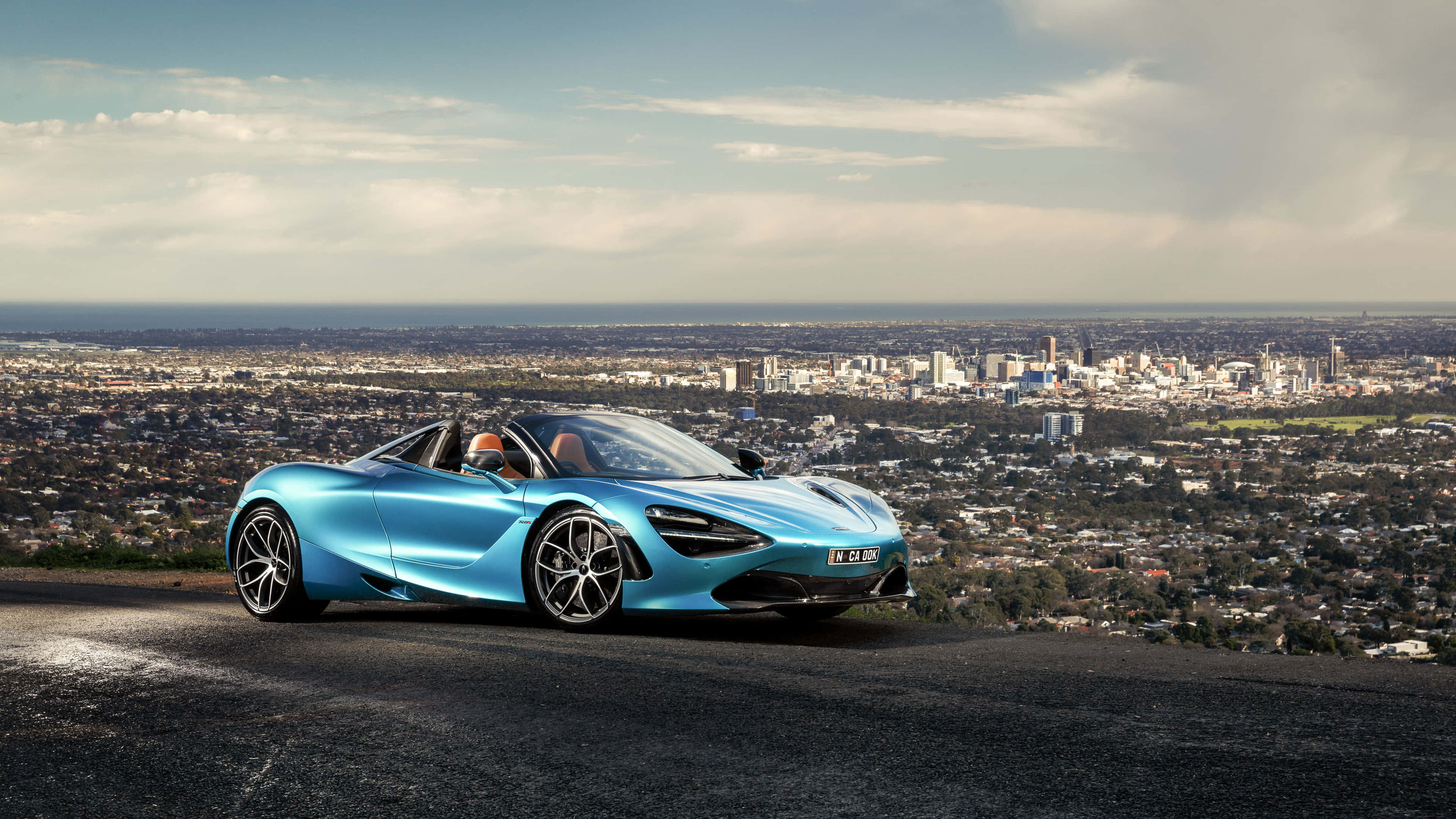 4K Mclaren 720s 桌布 壁紙 — 一架時尚的藍色敞篷跑車停泊在一個小山上，俯瞰著廣闊的城市景觀，天空部分多雲。