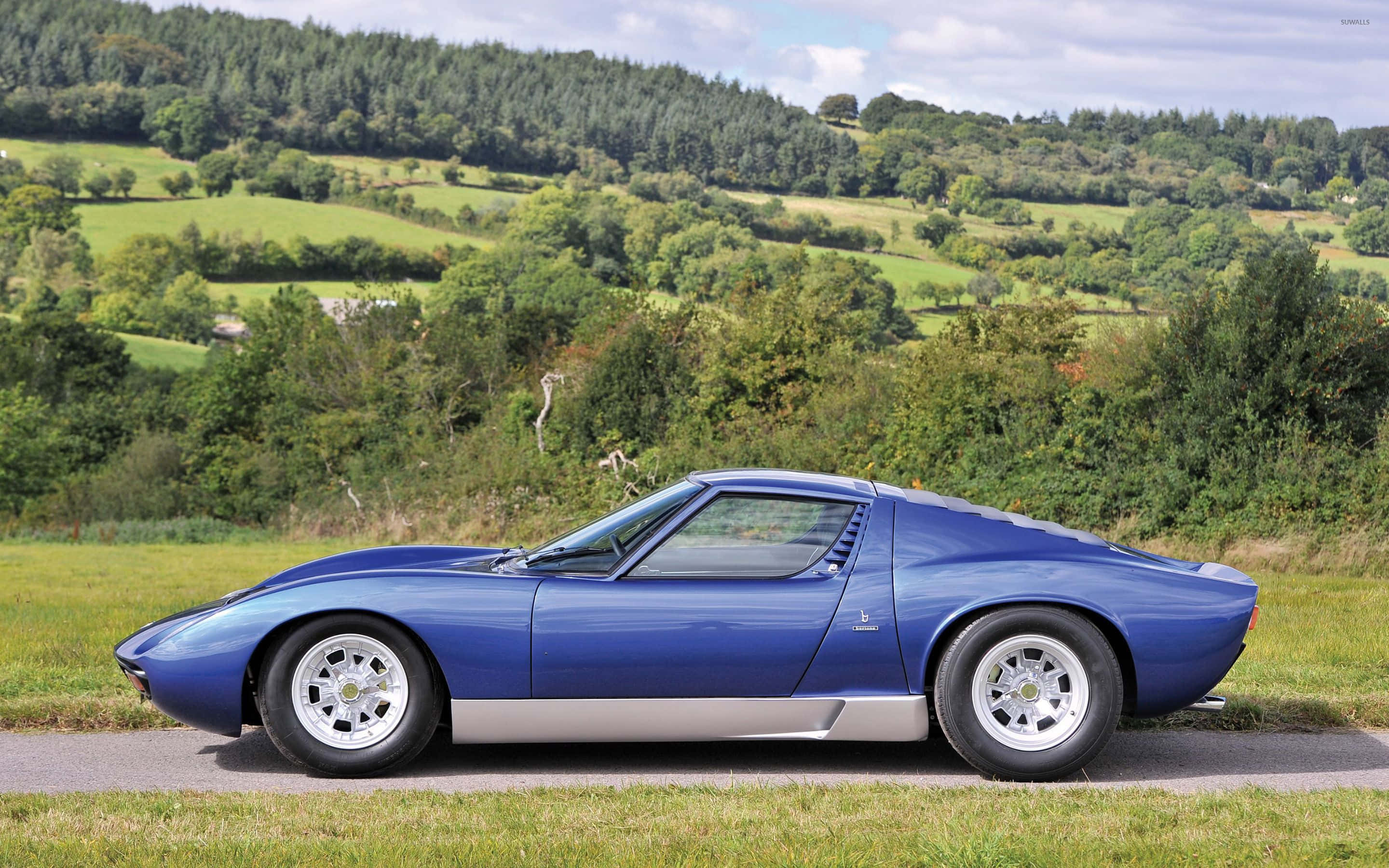 Lamborghini Miura 桌布 壁紙 — 一架時尚的藍色藍寶堅尼Miura跑車停泊在鄉村道路上，背景是起伏的綠色山丘和茂密的森林，天空部分多雲。