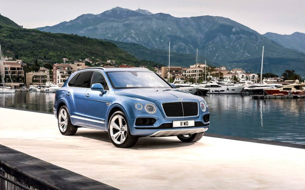 bentley HD 桌布 壁紙 — 一架時尚的藍色賓利 Bentayga SUV 停泊在海濱長廊上，背景有山脈和豪華遊艇。
