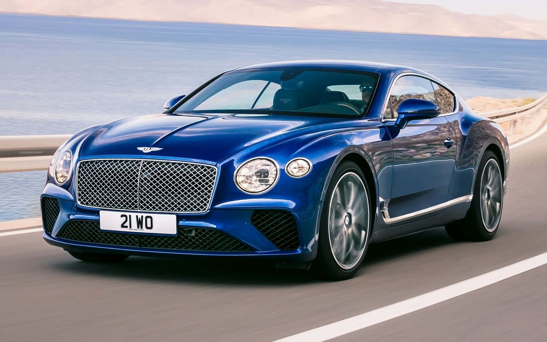 Best Bentley 桌布 壁紙 — 一架時尚的藍色賓利 Continental GT 沿著海岸公路行駛，背景是海洋和懸崖。