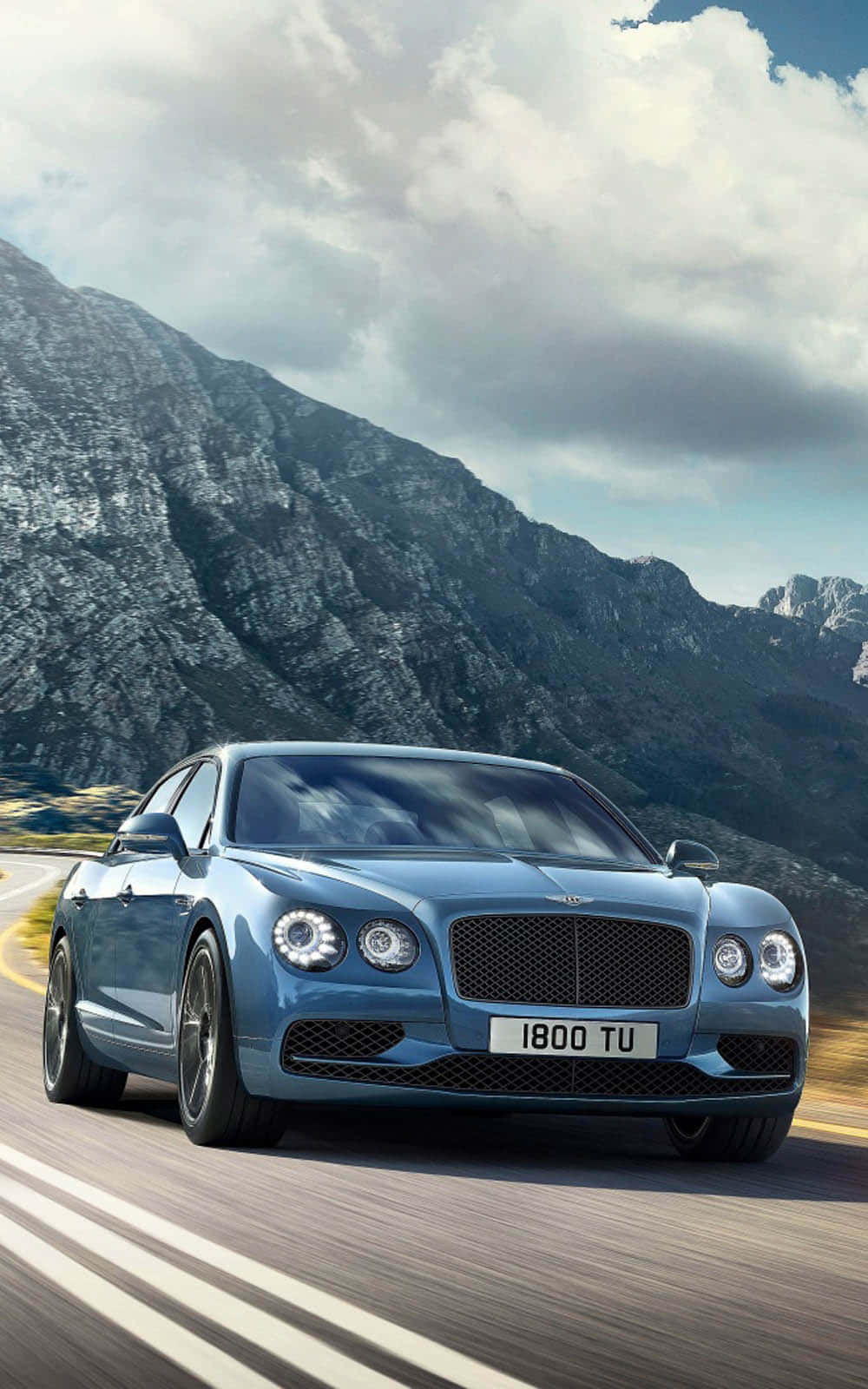 Bentley Flying Spur 桌布 壁紙 — 一架時尚的藍色賓利 Continental GT 在多雲的天空下沿著蜿蜒的山路飛馳。