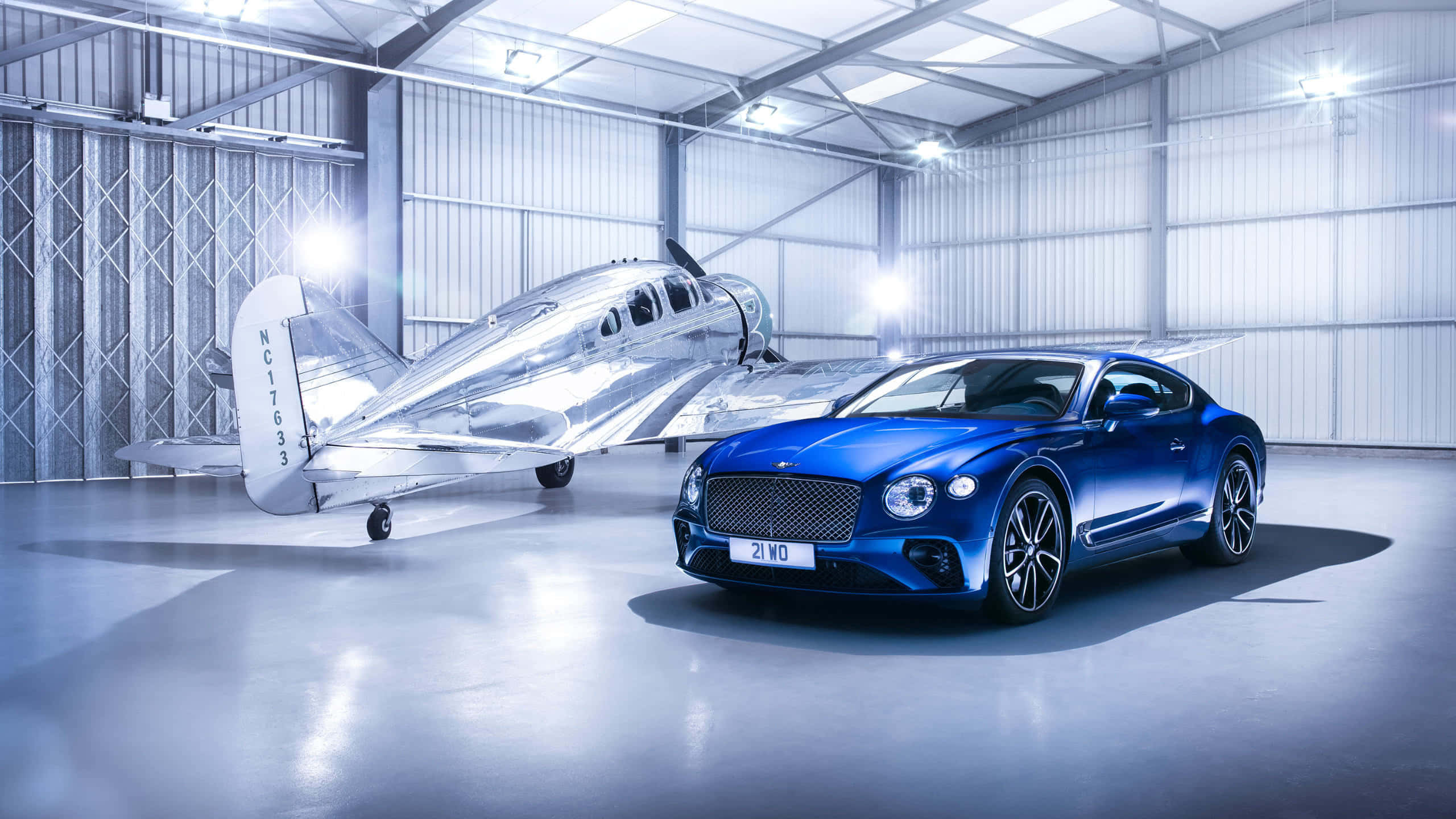 1440p Plane 桌布 壁紙 — 一架時尚的藍色賓利 Continental GT 停在一架銀色復古飛機旁邊，位於明亮的工業機庫內。