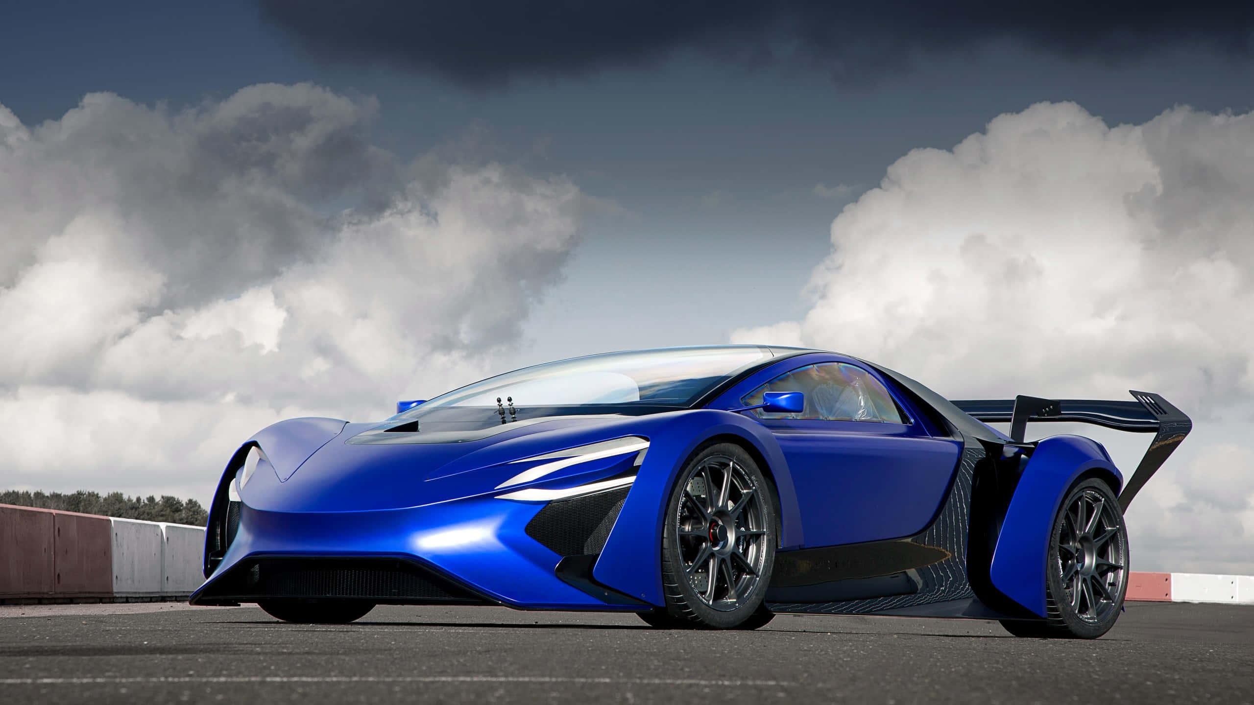 cars 桌布 壁紙 — 一架時尚的藍色超級跑車配備誇張的尾翼，在陰天下的賽道上蓄勢待發。