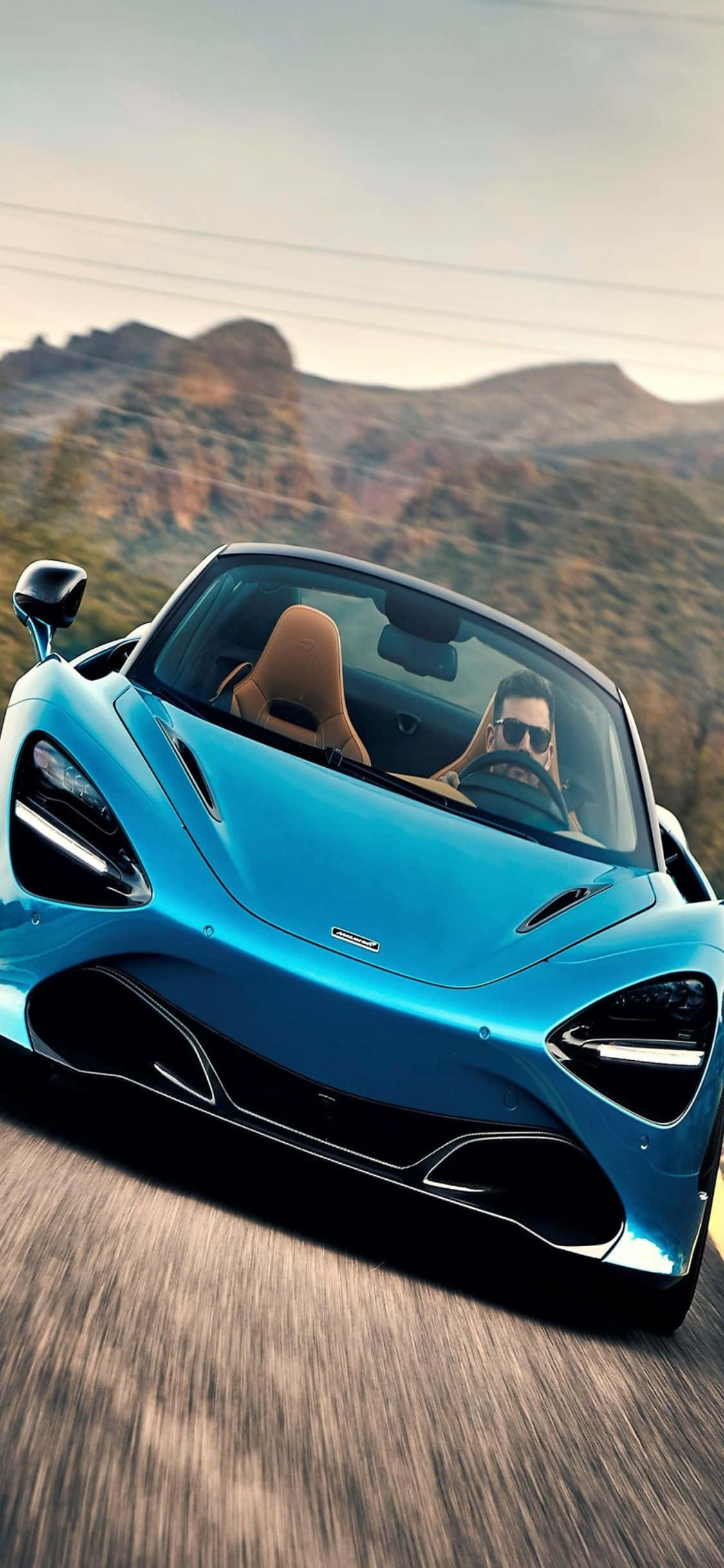iPhone X Mclaren 720s 桌布 壁紙 — 一架時尚的藍色跑車在沙漠峽谷中飛馳，司機戴著太陽眼鏡。