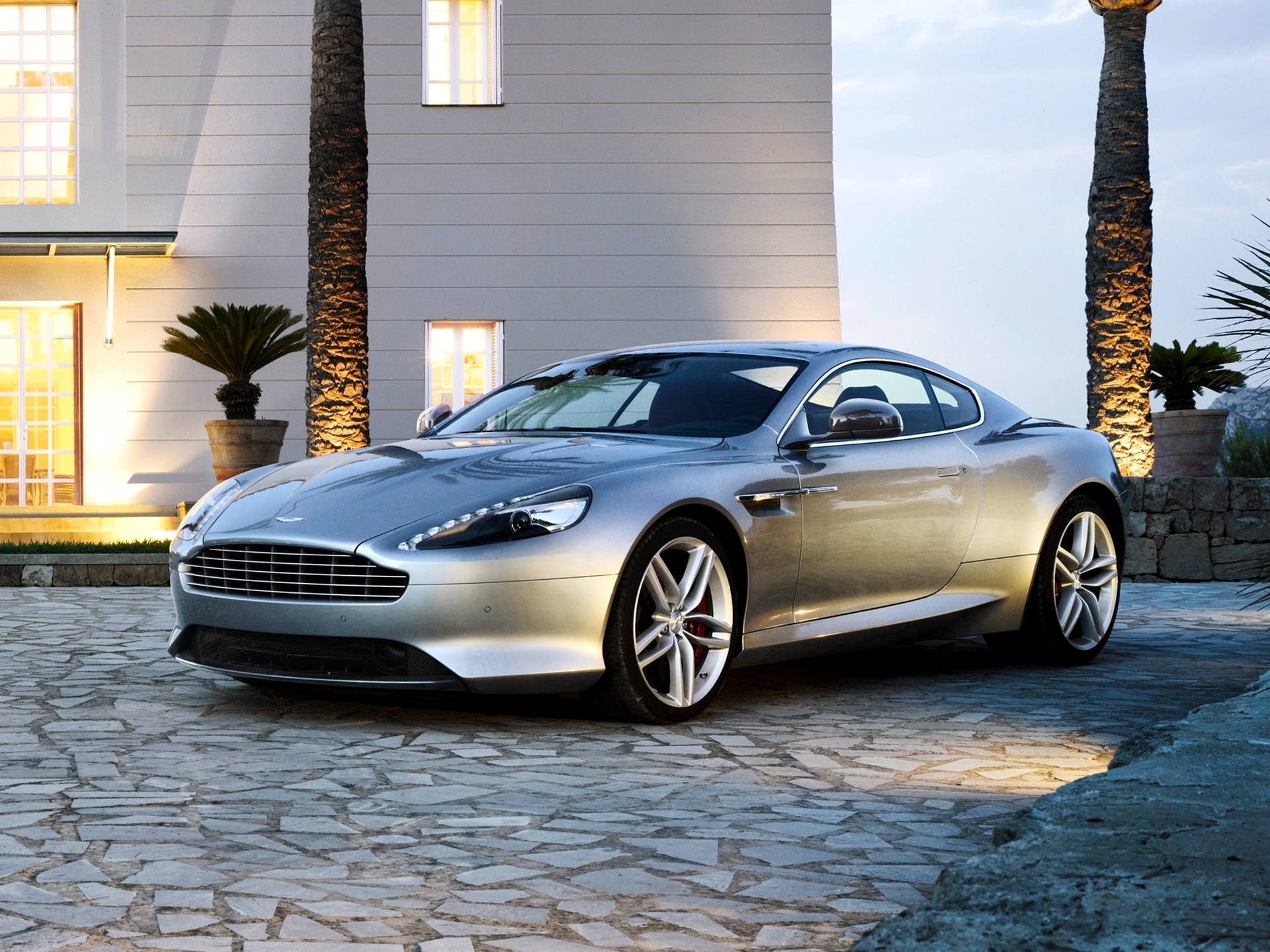 銀色 桌布 壁紙 — 一架時尚的銀色 Aston Martin 跑車在黃金時段於現代白色房屋前的石頭車道上閃閃發光。