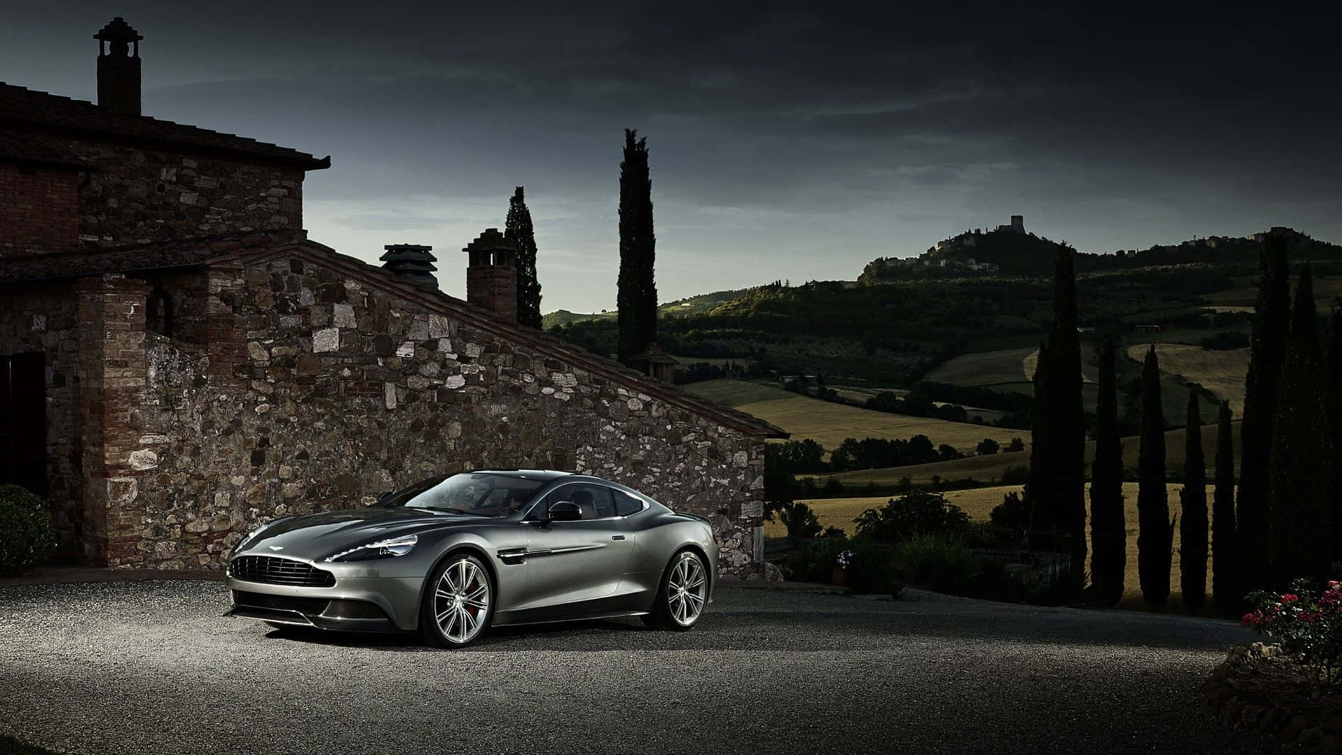 Aston Martin Vanquish 桌布 壁紙 — 一架時尚的銀色 Aston Martin 跑車停泊在碎石車道上，旁邊是一座質樸的石頭別墅，背景是陰鬱的暮色天空。