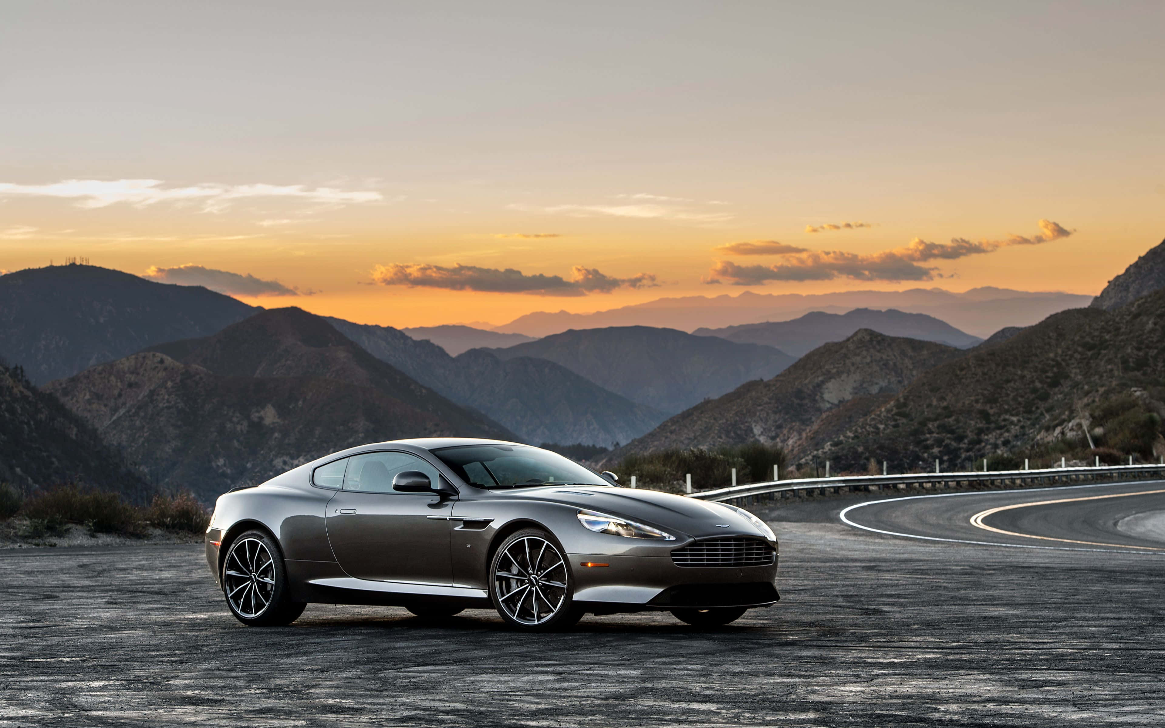 Aston Martin Db9 桌布 壁紙 — 一架時尚的銀色 Aston Martin 跑車停泊在蜿蜒的山路上，夕陽映照出橙色和紫色的天空。