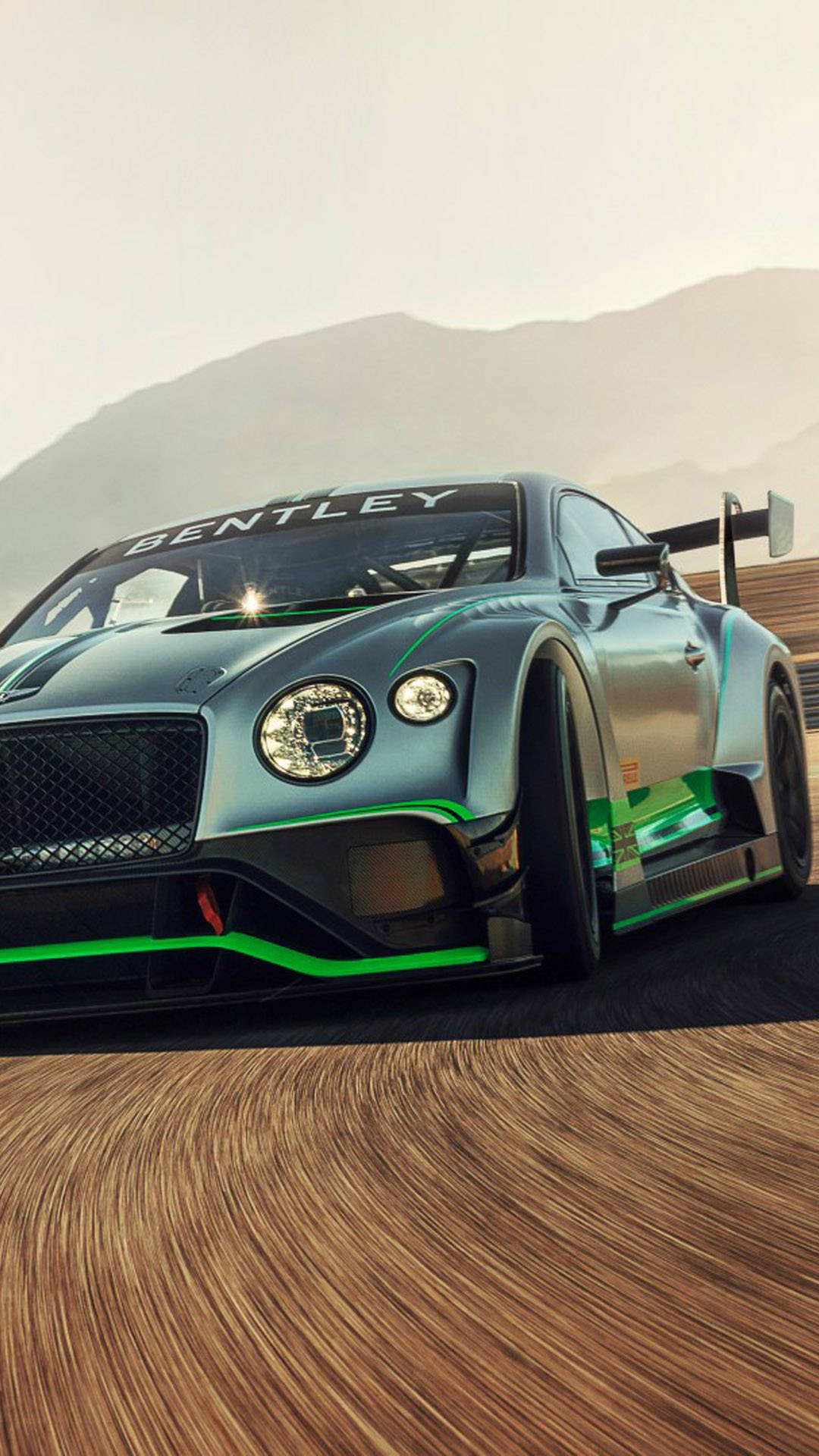 手機 桌布 壁紙 — 一架時尚的銀色 Bentley 賽車，帶有綠色點綴，沿著賽道疾馳，前景是模糊的木紋質感，背景是朦朧的山巒。