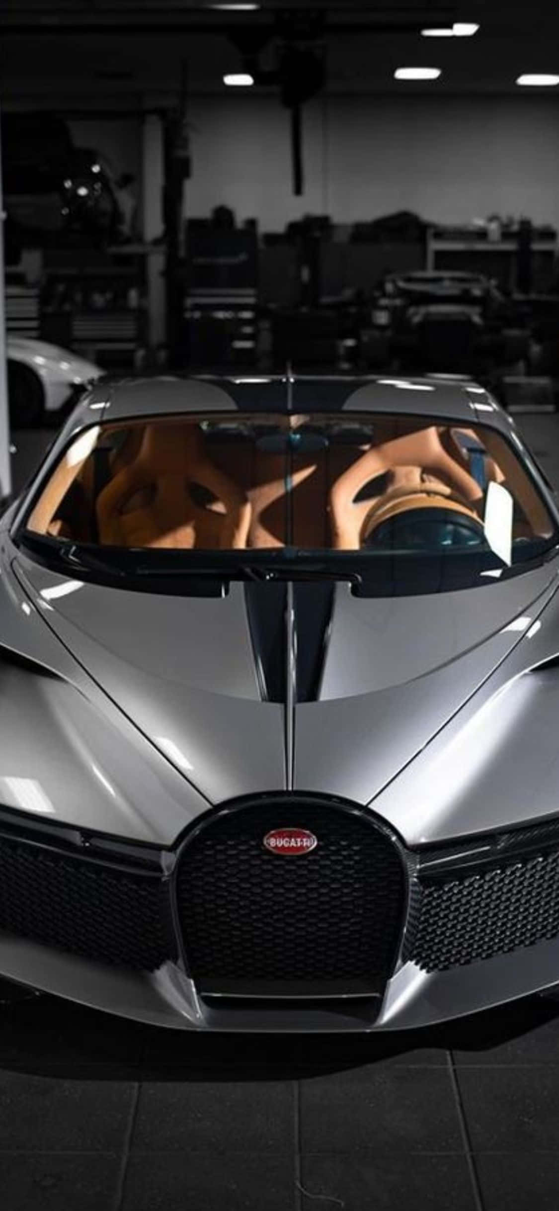 iPhone Xs Bugatti 桌布 壁紙 — 一架時尚的銀色 Bugatti 超級跑車配備棕色皮革座椅，停放在天花燈下光線昏暗的車庫中。