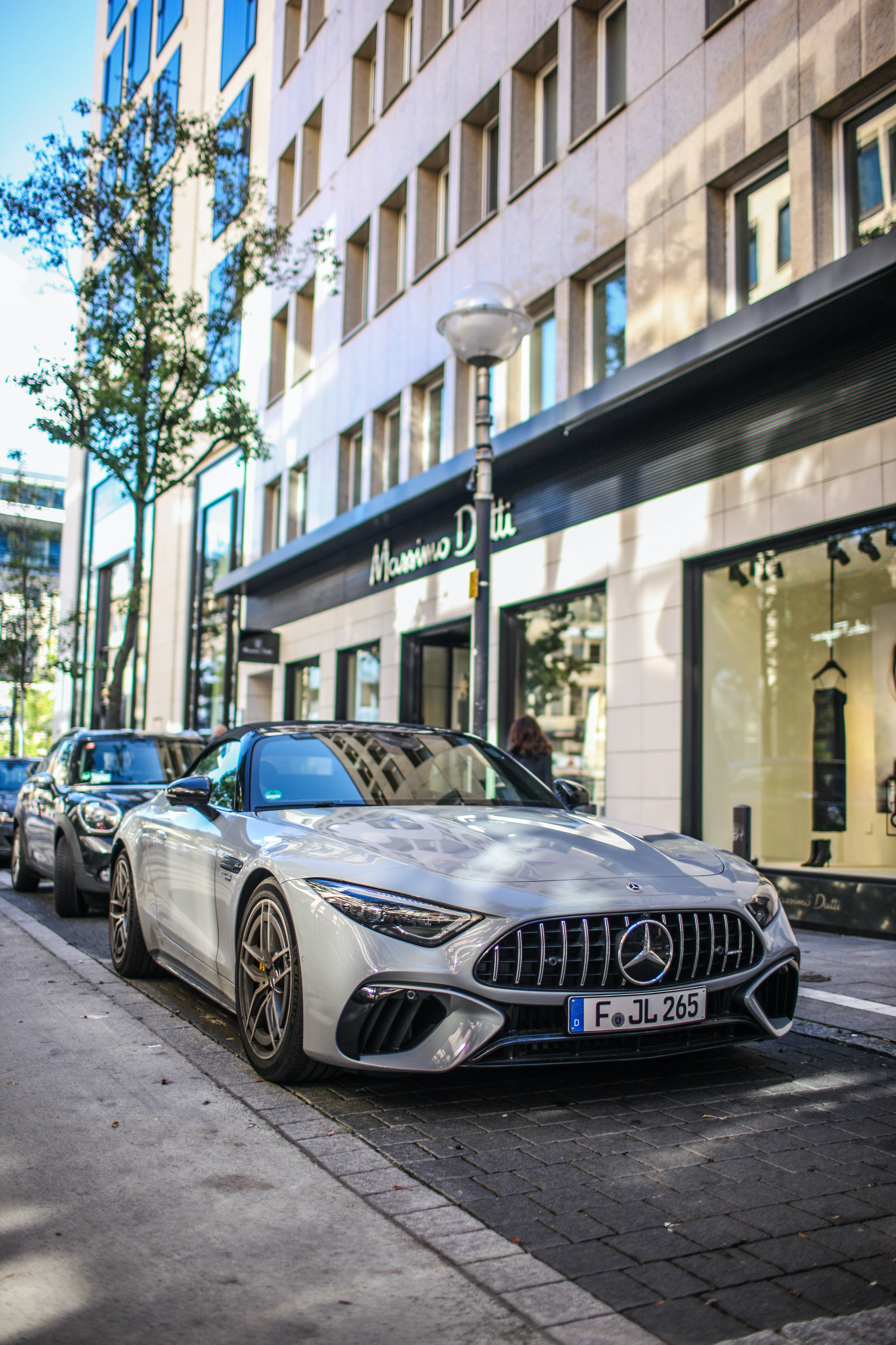 mercedes amg iPhone 桌布 壁紙 — 一架時尚的銀色 Mercedes-AMG GT 停泊在陽光明媚的城市街道上，旁邊是一家擁有現代建築和綠樹成蔭人行道的豪華精品店。