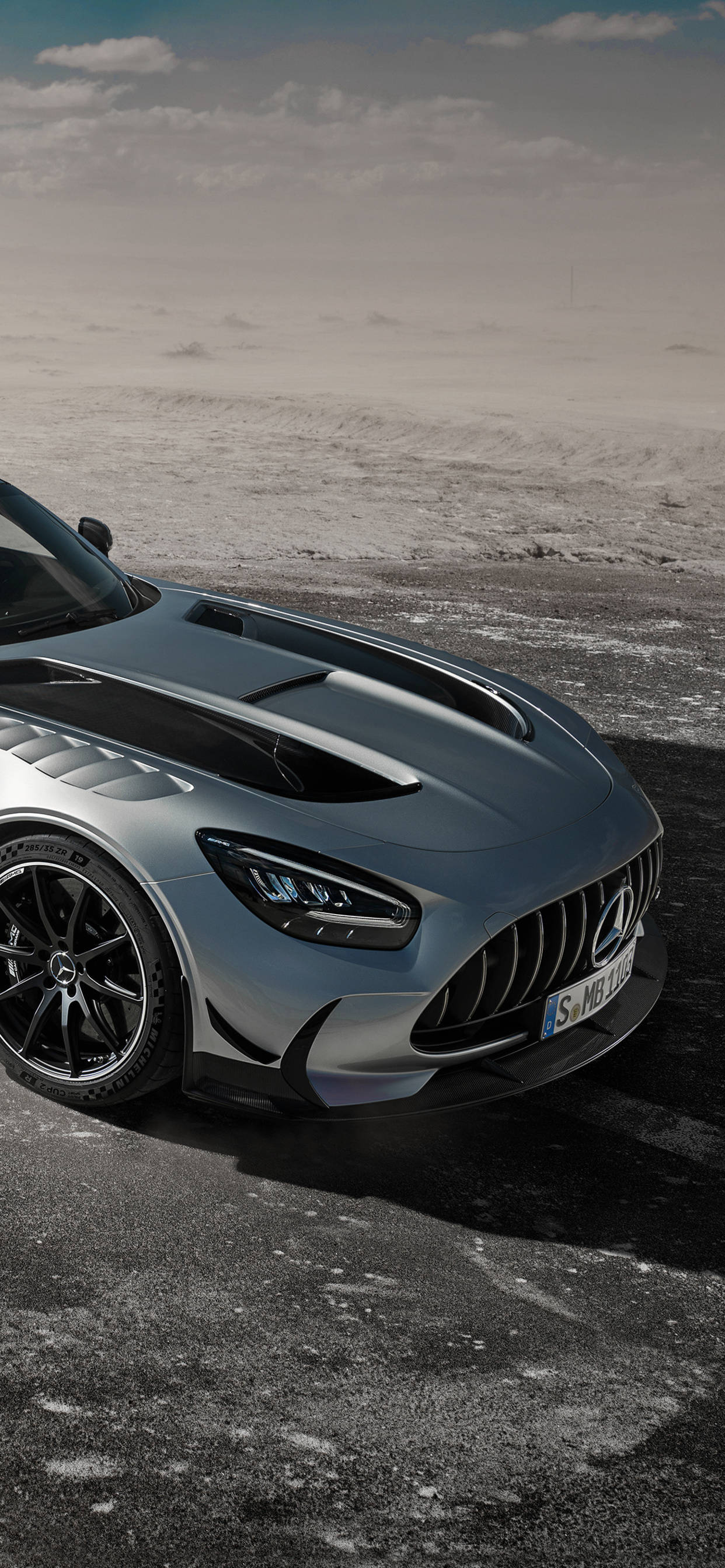 AMG iPhone 桌布 壁紙 — 一架時尚的銀色 Mercedes-AMG GT R 跑車停泊在龜裂的柏油路上，天空多雲。