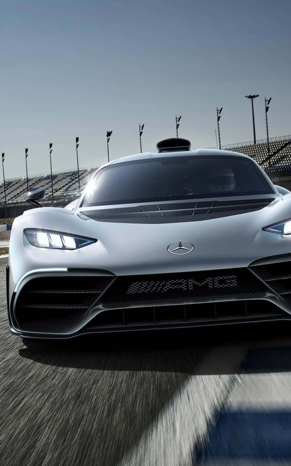 iPhone X Mercedes 桌布 壁紙 — 一架時尚的銀色 Mercedes-AMG 賽車在賽道上疾馳，背景有模糊的動感和體育場燈光。