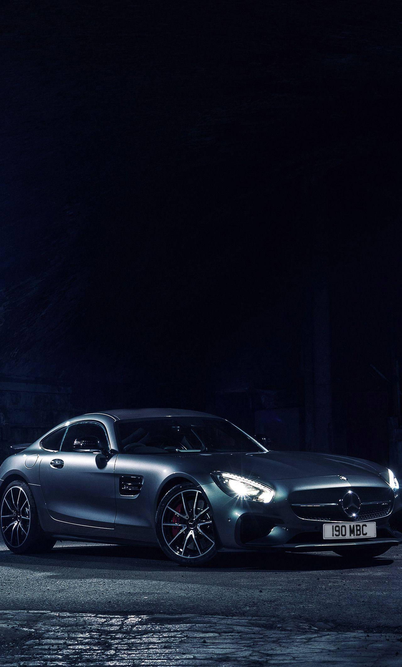 賓士 iPhone 桌布 壁紙 — 一架時尚的銀色 Mercedes-AMG GT 跑車在夜晚的黑暗鵝卵石街道上，在自己的車頭燈下發光。