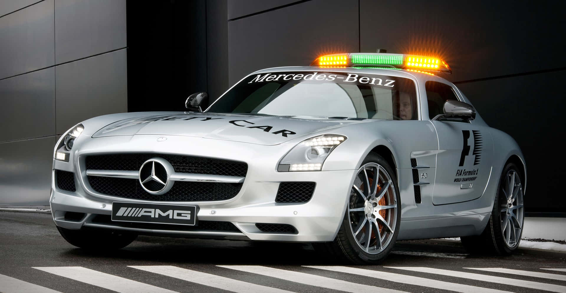Safety Car 桌布 壁紙 — 一架時尚的銀色 Mercedes-AMG SLS GT1 安全車，閃爍著燈光，停放在斑馬線上，背景是一座深色現代建築。