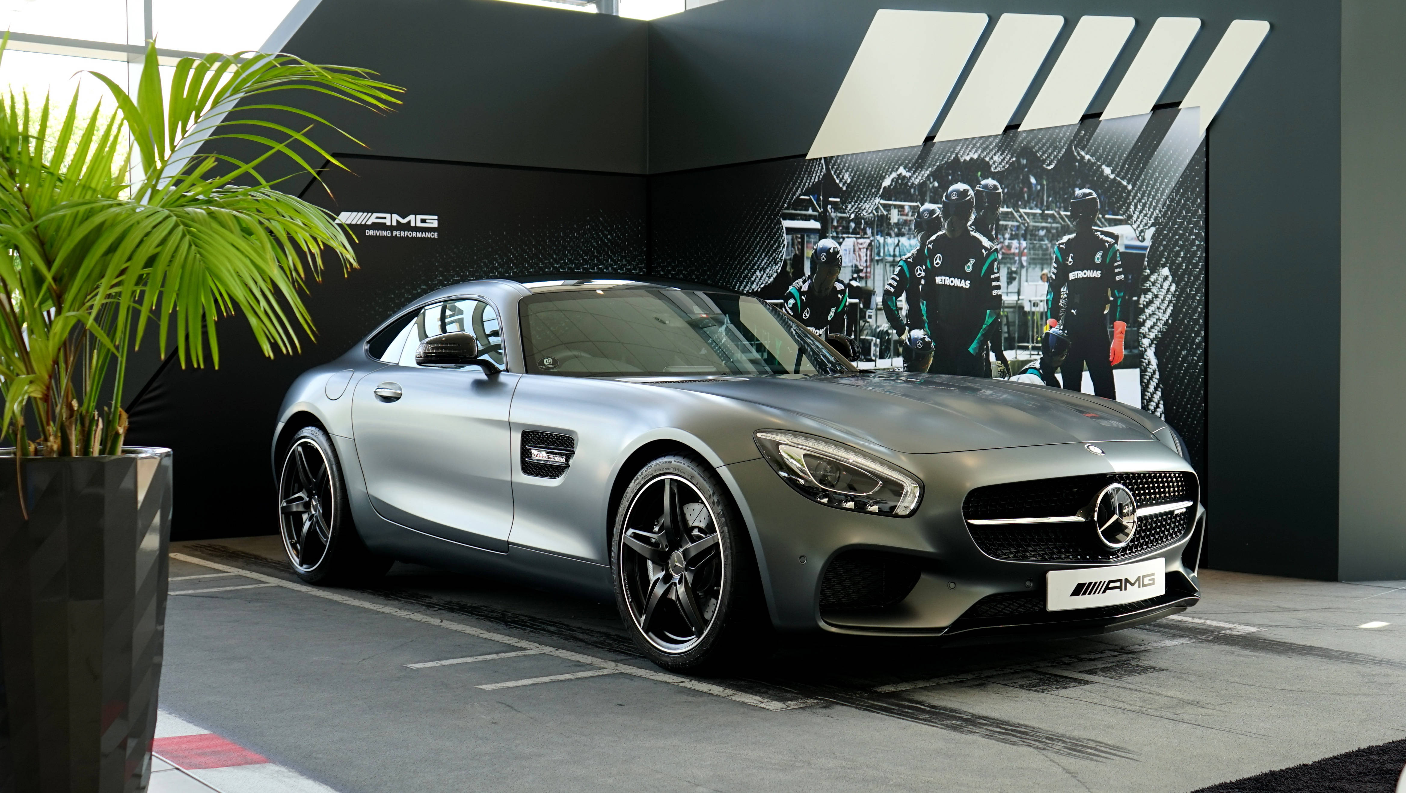amg iPhone 桌布 壁紙 — 一架時尚的銀色 Mercedes-AMG GT 跑車在室內展示，旁邊有一棵盆栽棕櫚樹和賽車主題牆面圖案。