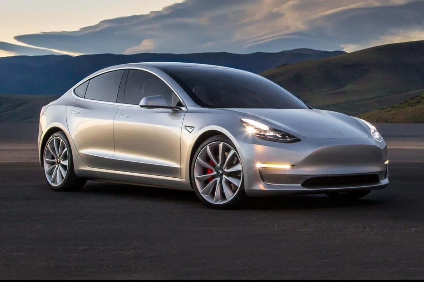 Tesla Model 3 桌布 壁紙 — 一架時尚的銀色 Tesla Model 3 停泊在黑暗的道路上，背景有山脈和多雲的天空。