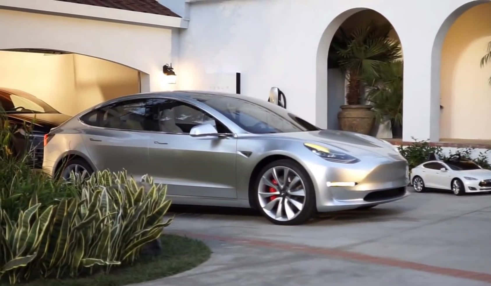 Tesla Model 3 桌布 壁紙 — 一架時尚的銀色 Tesla Model 3 停放在白色灰泥房屋前，房屋有拱形門口和盆栽棕櫚樹。
