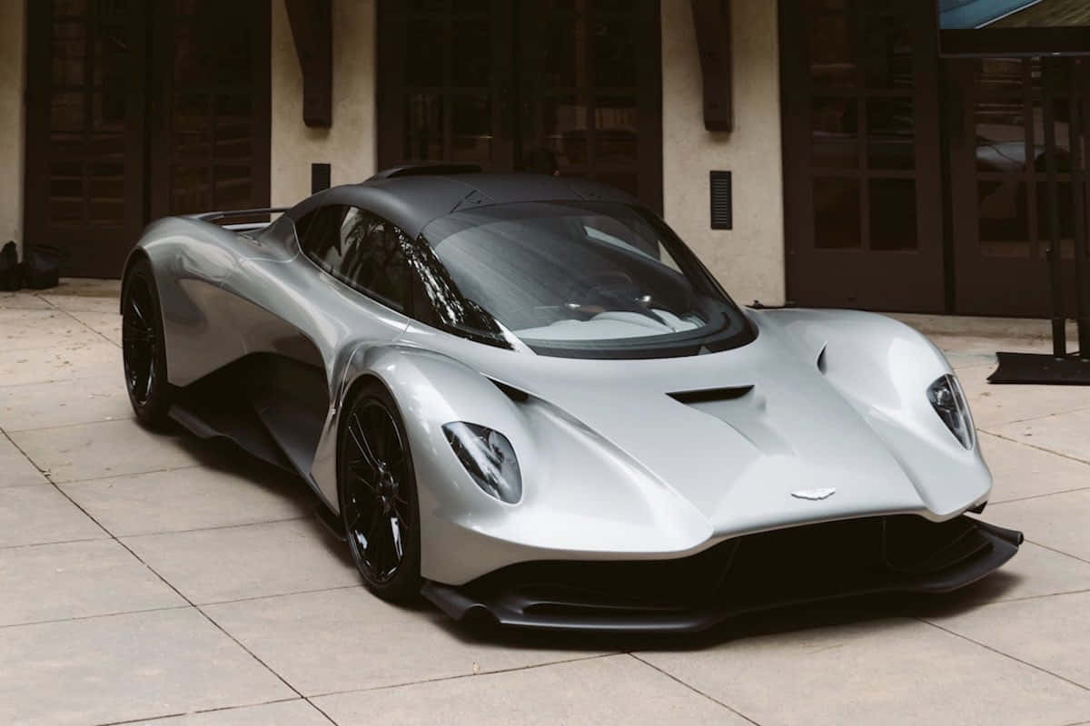 奧斯頓馬丁 桌布 壁紙 — 一架時尚的銀色Aston Martin Valkyrie超跑停泊在瓷磚庭院，背景有建築物。