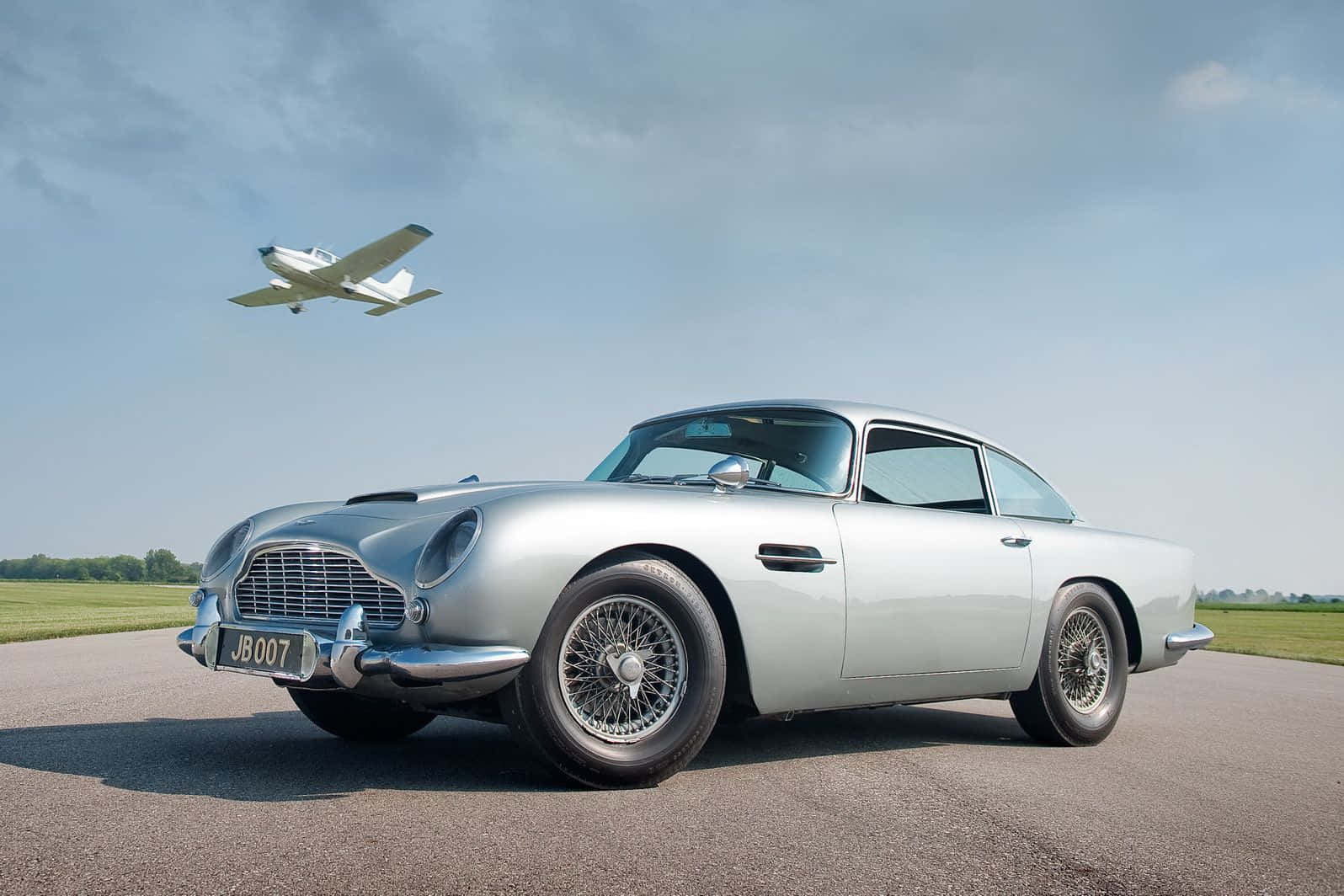 Aston Martin Db5 桌布 壁紙 — 一架時尚的銀色Aston Martin DB5停在飛機跑道上，一架白色小飛機在部分多雲的天空下飛過頭頂。