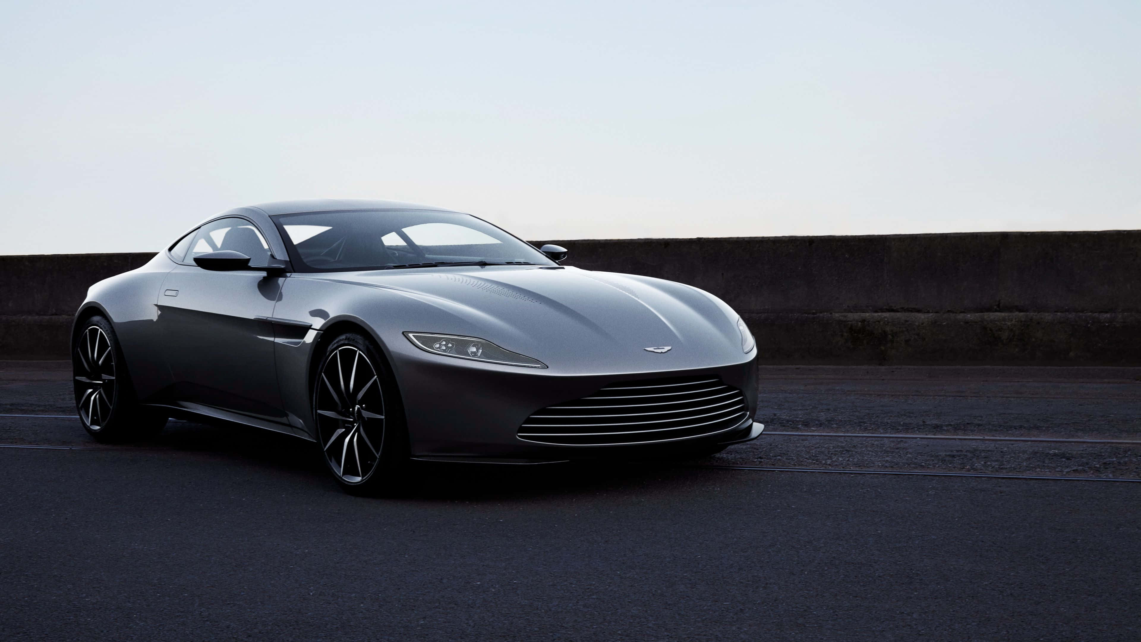 奧斯頓馬丁 桌布 壁紙 — 一架時尚的銀色Aston Martin跑車停泊在昏暗的瀝青路上，旁邊是混凝土護欄，天空蒼白。