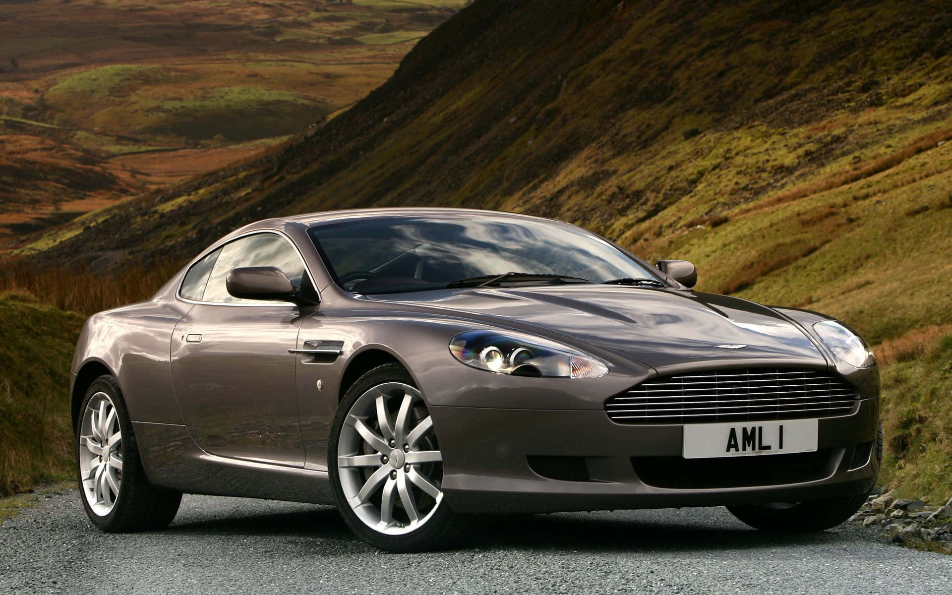 Aston Martin Db9 桌布 壁紙 — 一架時尚的銀色Aston Martin DB9雙門跑車停泊在碎石路上，背景是起伏的綠色山丘。